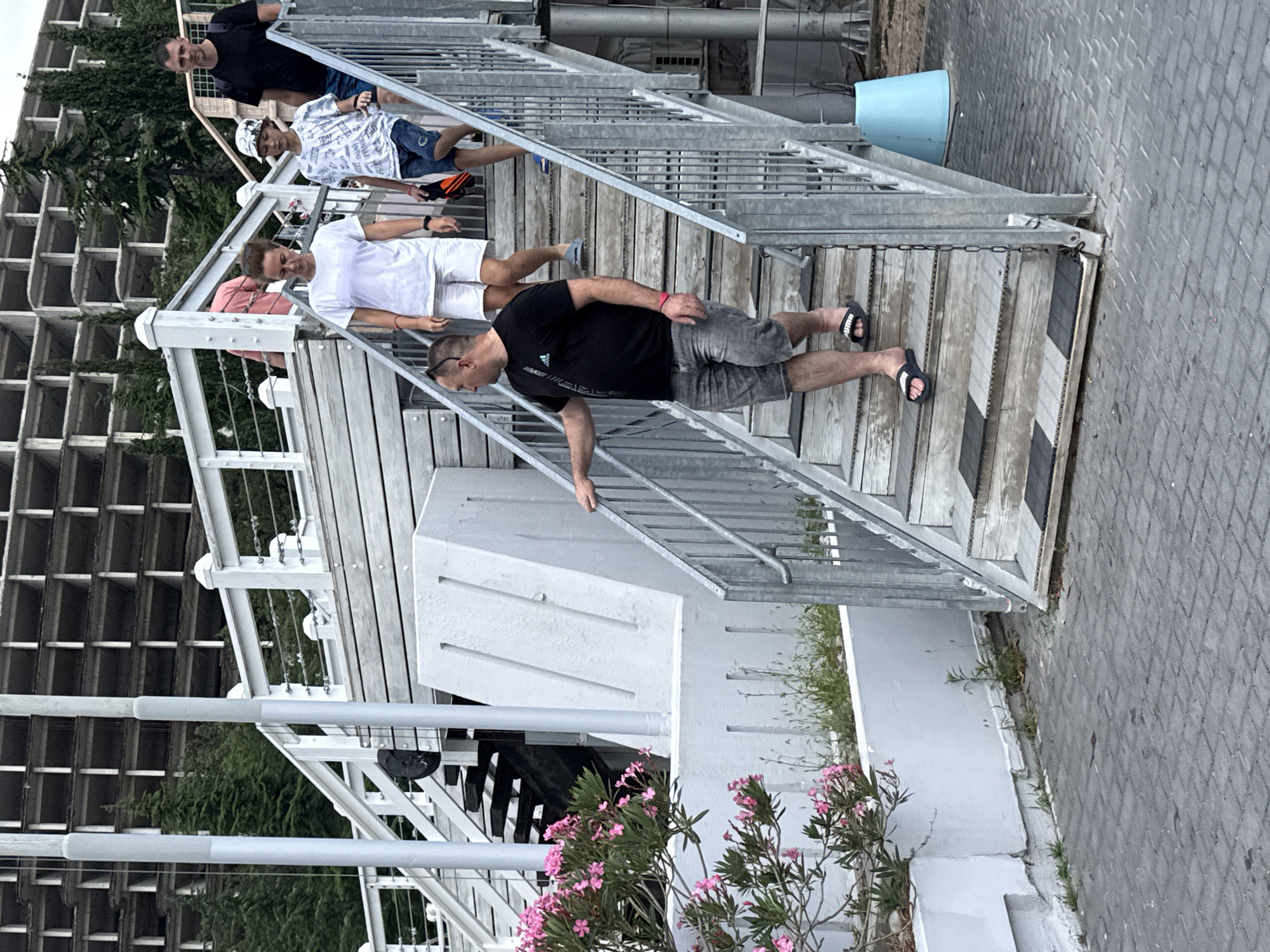The image shows an outdoor staircase with several people descending. A man in a black t-shirt and gray shorts is in the foreground, wearing black flip-flops. He is looking to his left. Behind him, a woman in a white t-shirt and white shorts, and a boy in a patterned t-shirt and blue shorts are also going down the stairs. Further up the stairs, a man in a black t-shirt and dark shorts is visible. The staircase is made of wood and metal. In the background, a multi-story building with a concrete frame is under construction. Pink flowers are blooming in a planter to the left of the stairs. The ground is paved with grey cobblestones. The scene appears to be in a city, likely a resort area given the architecture and landscaping. The location is identified as Yalta, Ukraine.