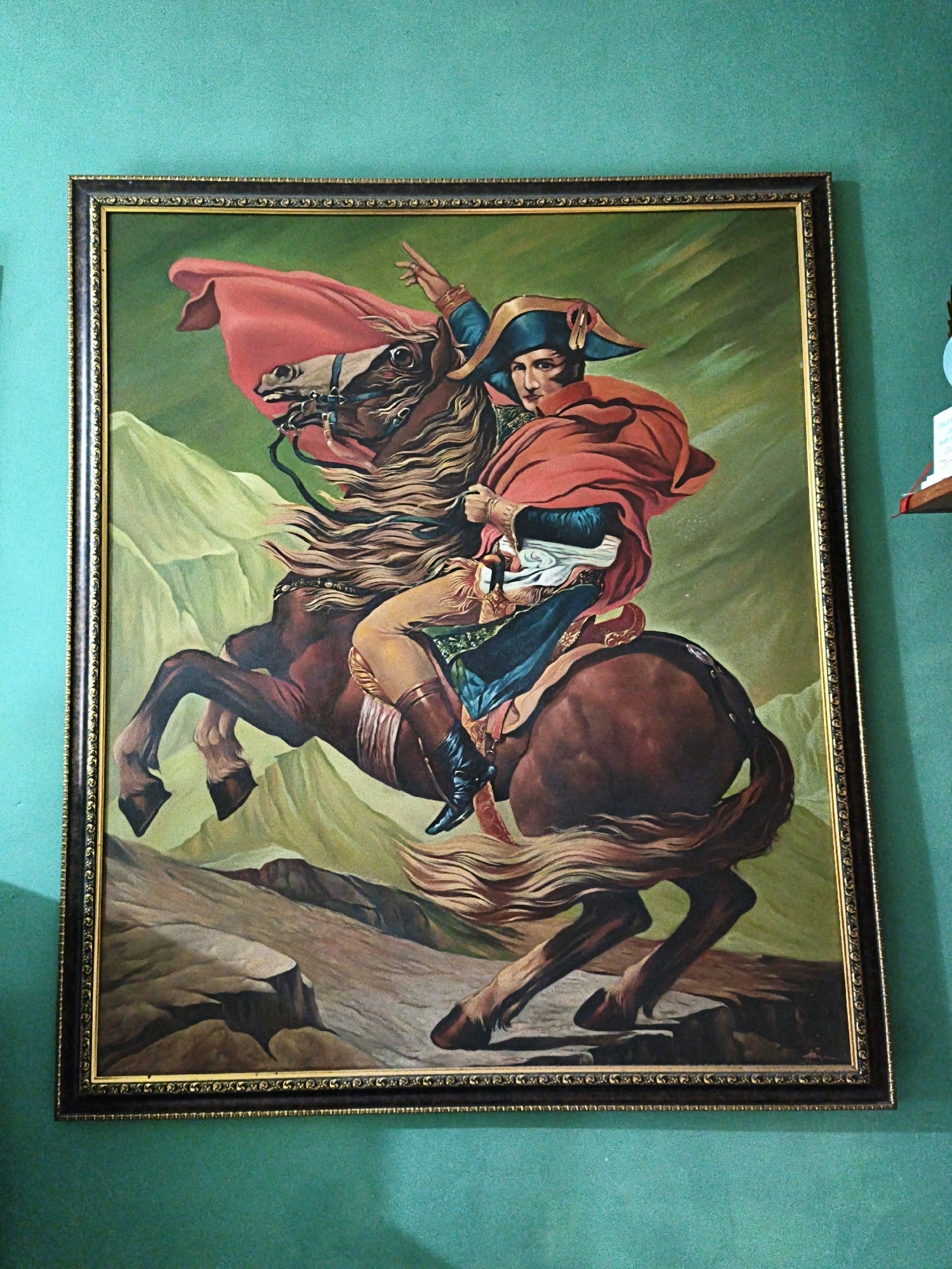 A framed painting of Napoleon Bonaparte on horseback is mounted on a teal wall. The painting depicts Napoleon in a red cape and blue and gold military attire, with a bicorne hat. He is shown with his left arm raised, pointing forward, and his right hand on the reins. The horse is brown and appears to be galloping across a mountainous landscape with green slopes and rocky terrain. The painting has a decorative gold and dark brown frame. A small wooden shelf with books is visible on the wall to the right of the painting. The image does not provide any information about the location in the city of Pabelan, Indonesia.