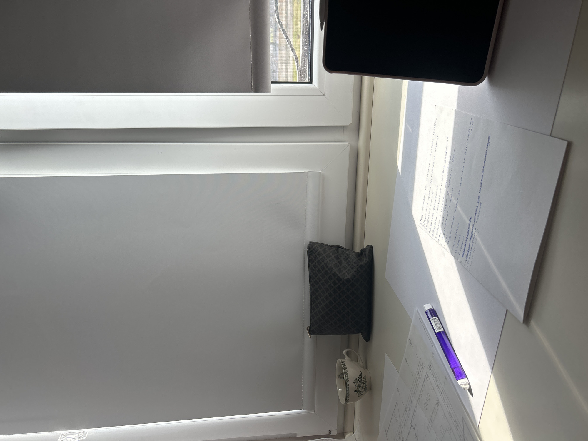 A high-angle shot captures a partially obscured window with a closed, light-colored blind. A dark, rectangular makeup bag rests on the windowsill near a small, porcelain cup with floral designs. Adjacent to these items, loose sheets of paper covered in handwritten notes and diagrams are visible. A purple pen lies atop one of the papers. The top edge of a black electronic device, likely a tablet or phone, is also visible at the top right of the image.
The handwritten text on the papers seems to be in Bulgarian, potentially related to construction or architectural design elements. The phrase "Алуминиев профил за монтаж на парапет" (Aluminum profile for parapet mounting) and mentions of glass ("стъкло") and wood ("Дървена") are discernible.
The scene suggests a workspace, possibly a home office or study area, where someone is working on design or technical documentation. While the setting and contents are visible, there are no people present in the image to provide additional context about their actions or interactions within the scene. The city of Plovdiv, Bulgaria is not visibly represented in the image.