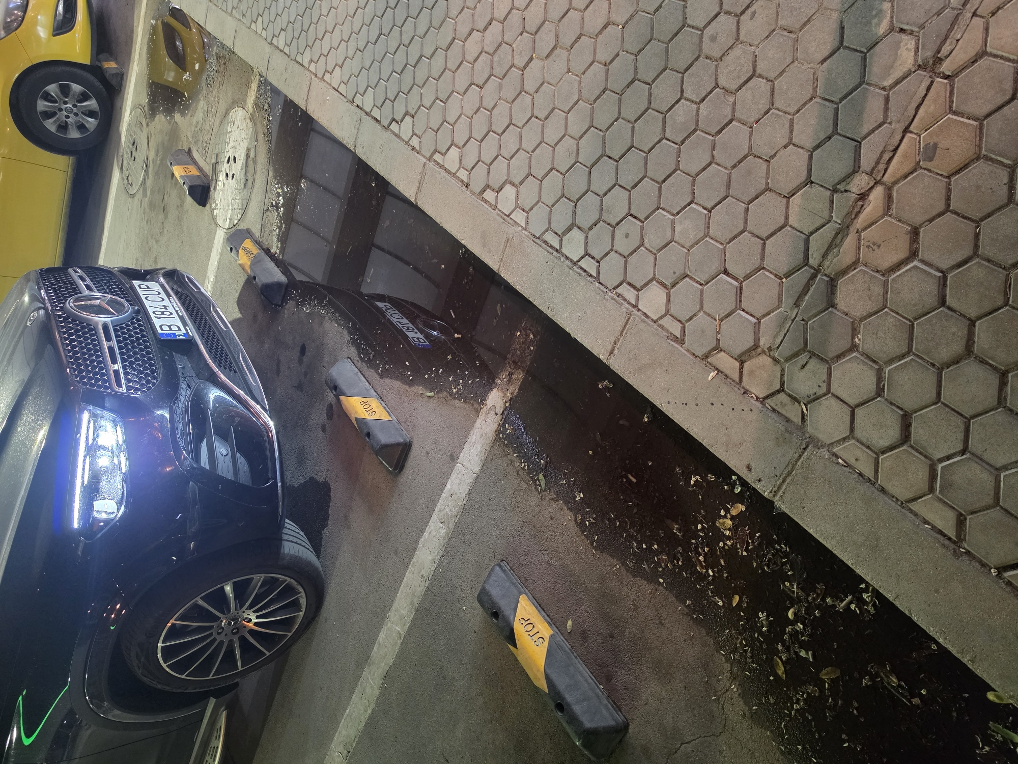 The image shows a black Mercedes-Benz GLB 200, license plate B 184 CUP, parked on asphalt. The car's headlights are on, illuminating the area. Next to the car, there is a wet patch on the ground, possibly from rain or a leak. Two black and yellow speed bumps with the word "STOP" are visible on the asphalt. A yellow car is parked behind the Mercedes. The scene appears to be a parking lot, possibly in Varna, Bulgaria, given the license plate format. The ground is covered with hexagonal paving stones on the right side of the image. The overall lighting suggests it is nighttime. There are no people visible in the image.