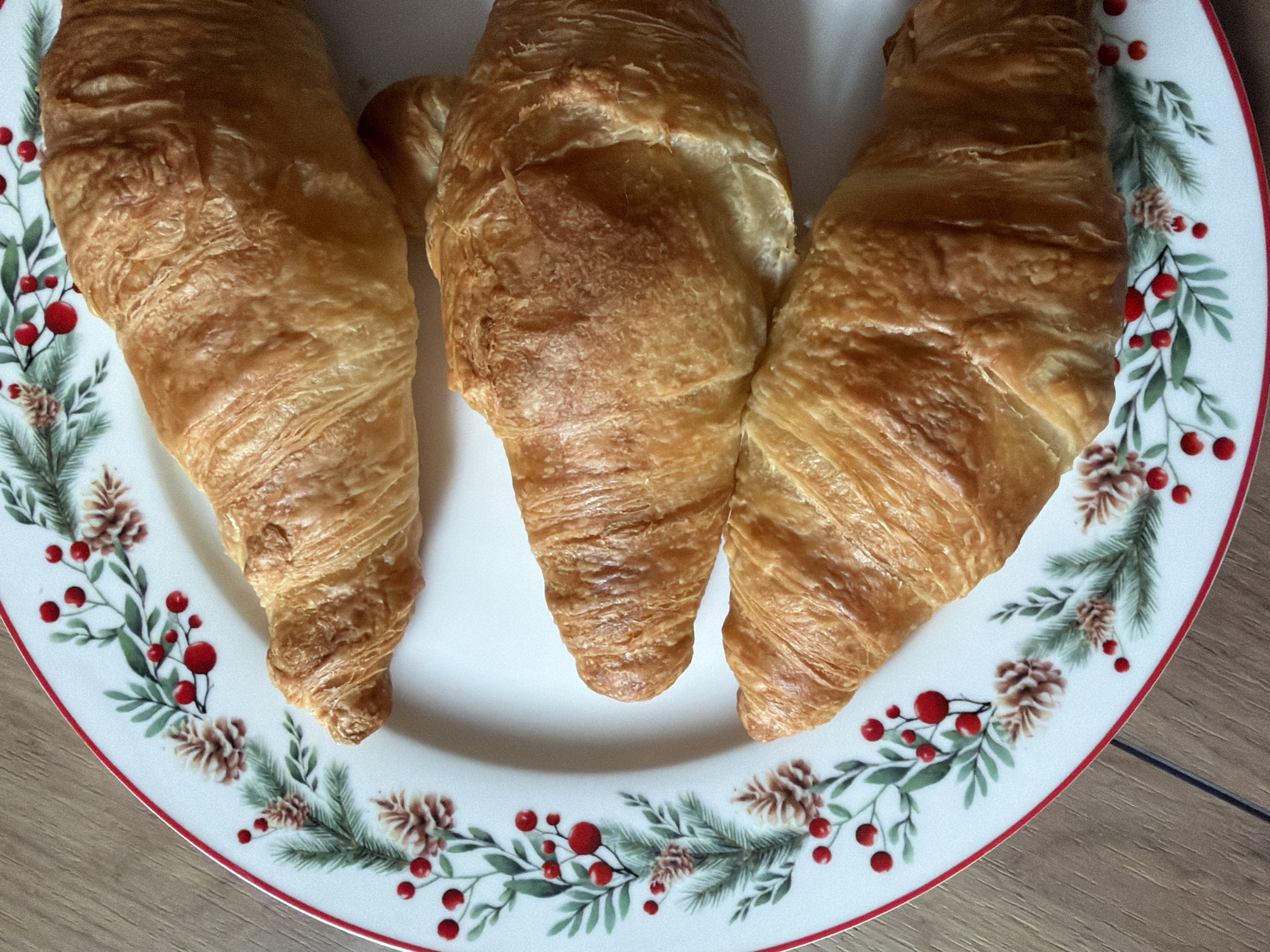 Three croissants are arranged on a white plate with a decorative border of green foliage, red berries, and brown pine cones. The plate sits on a wooden surface with light brown hues. The image does not contain any people or any information to determine that the picture was taken in Kazan, Russia.
