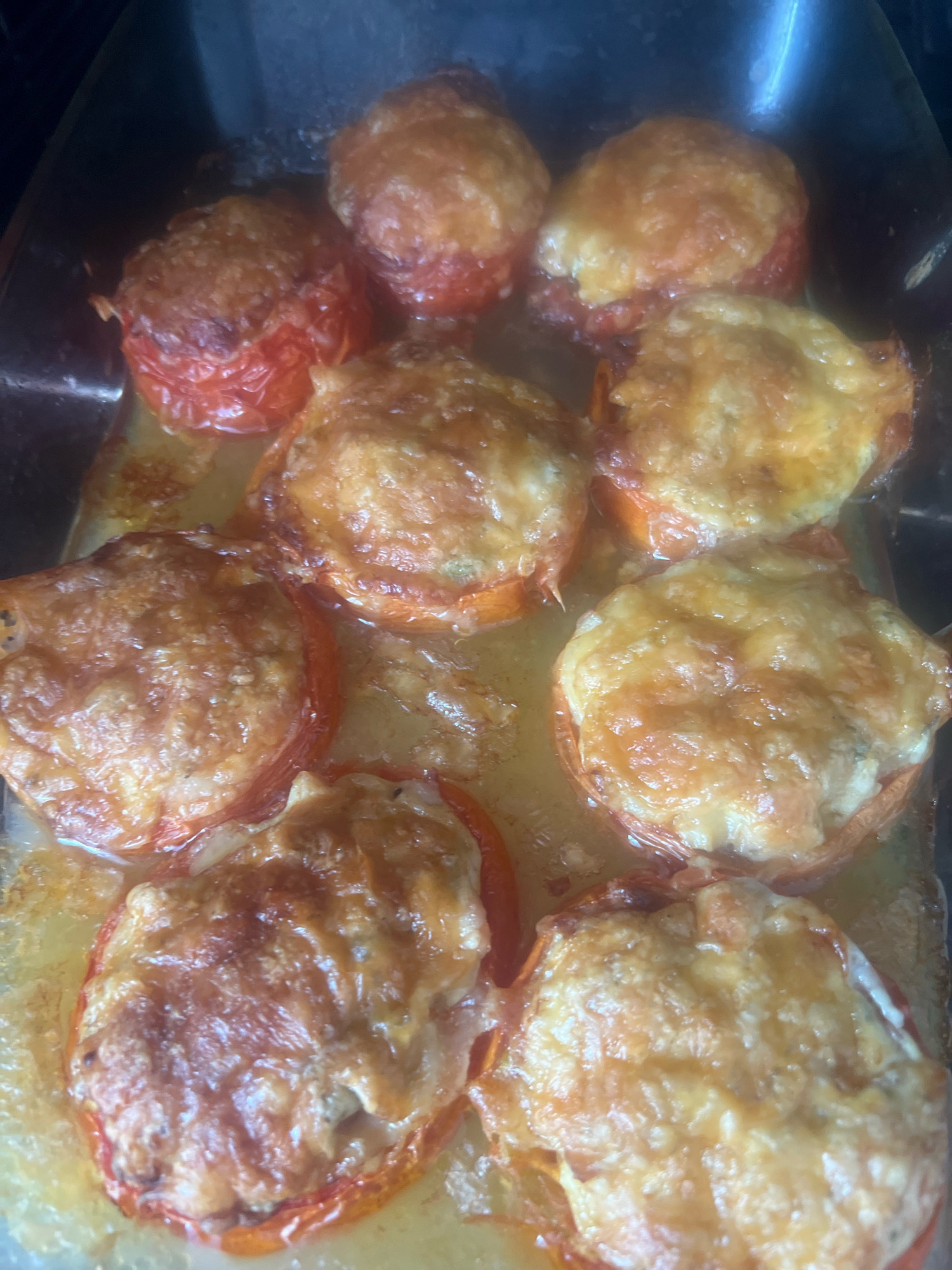 This is a top-down, close-up shot of baked tomatoes with cheese topping, arranged in a metal baking dish. The tomatoes appear to be whole or halved, with the filling and melted, golden-brown cheese covering the tops. Some of the tomatoes have slightly browned edges, indicating they have been roasted or baked to a crisp. The dish is filled with some liquid, likely juices from the tomatoes and any other ingredients in the filling. The lighting is warm and natural, highlighting the texture of the cheese and tomatoes. The image does not contain any identifiable landmarks or elements that would suggest a specific location in Otradnoye, Russia, or any city for that matter.