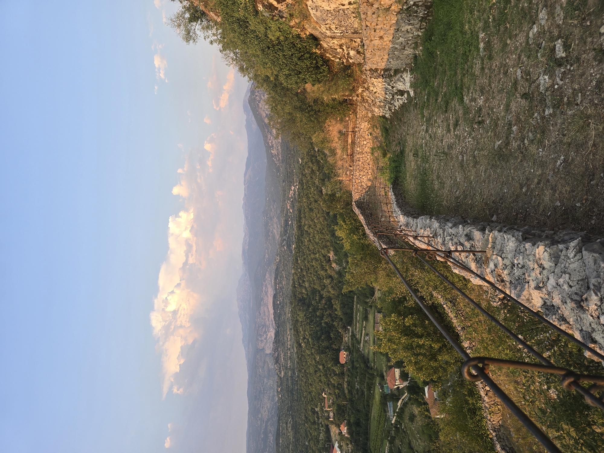 The photograph captures a landscape scene with a pathway leading along a stone wall with a metal railing on the left. The pathway is partially overgrown with grass and rocks. Beyond the pathway, a valley is visible, dotted with houses and lush green vegetation. In the distance, rolling hills and mountains form the horizon under a pale blue sky with scattered, soft clouds. The lighting suggests it is late afternoon or early evening, with the sun casting a warm glow on the stone wall and the distant landscape. The location is identified as Gornja Vrbica, Montenegro. There are no people or animals visible in the image. The scene is tranquil and offers a panoramic view of a rural mountainous area.