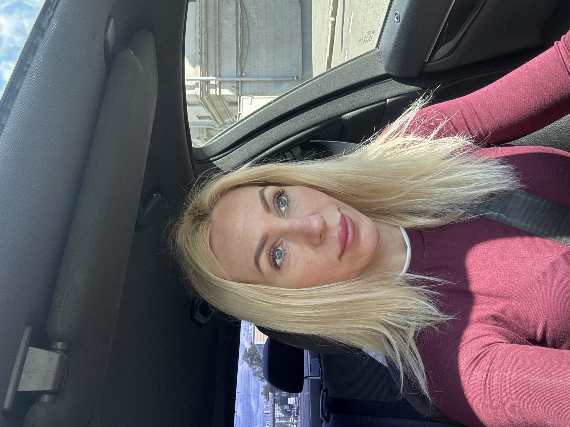 A blonde woman is in the driver's seat of a car. She is wearing a burgundy long-sleeved shirt. The car interior is dark gray. Through the windshield and side window, a city street is visible with buildings and traffic. The sun is shining, casting shadows inside the car. The background outside shows buildings with classical architecture, suggesting a historical part of Saint Petersburg, Russia. The sky is partly cloudy.