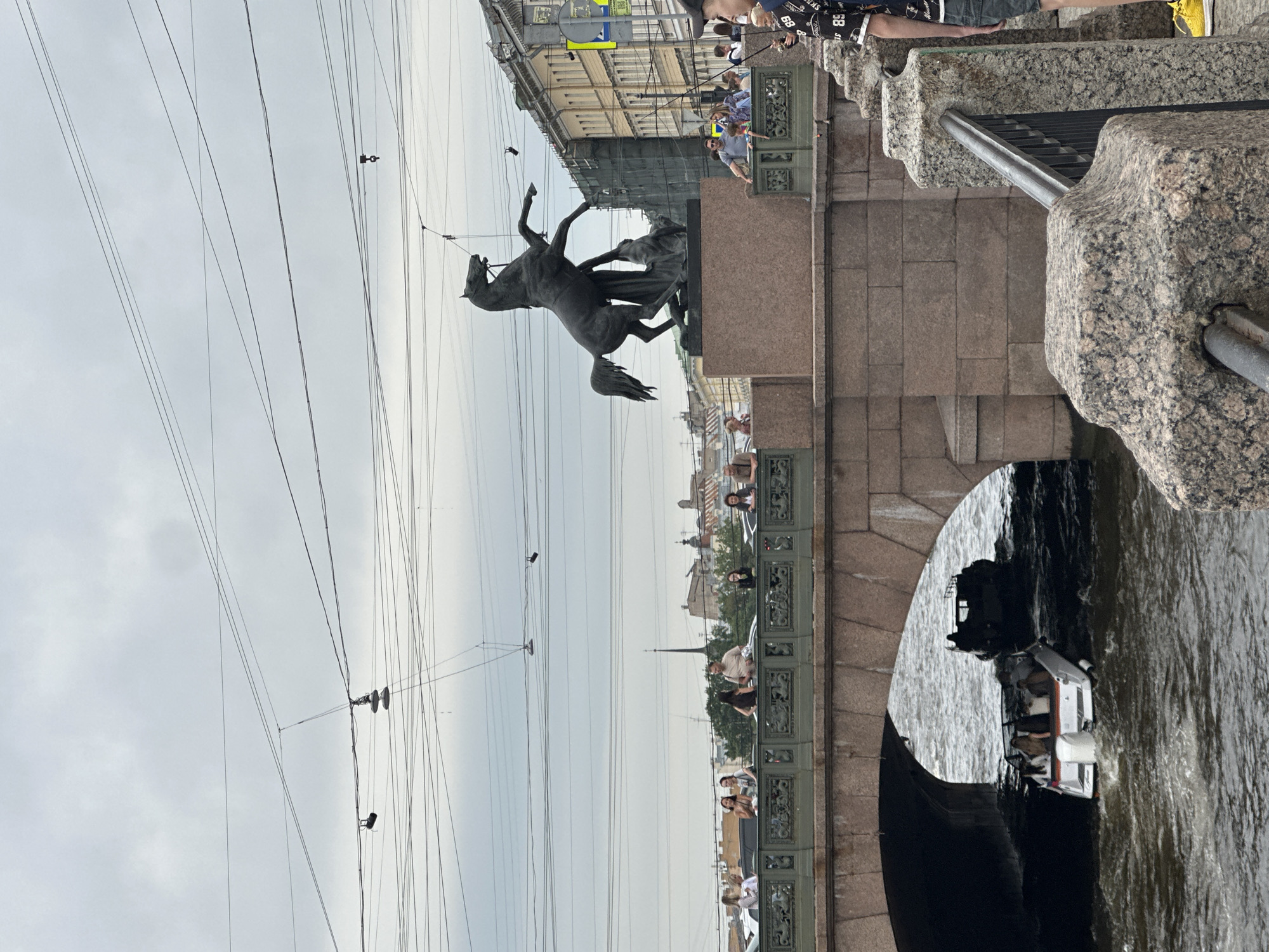 This image depicts the Anichkov Bridge in Saint Petersburg, Russia. The iconic equestrian statue "Horse Tamer" by Pyotr Klodt stands prominently on the bridge. Several people are visible on the bridge's railing, some looking out over the water, while others appear to be posing for photos. Below the bridge, a small boat navigates the Fontanka River. The sky is overcast, and there are numerous overhead wires, likely for trolleybuses or other urban transport. The architecture of the surrounding buildings, characteristic of Saint Petersburg, is visible in the background. The scene captures a typical day on this historic bridge, with both tourists and locals enjoying the view and the city's atmosphere.