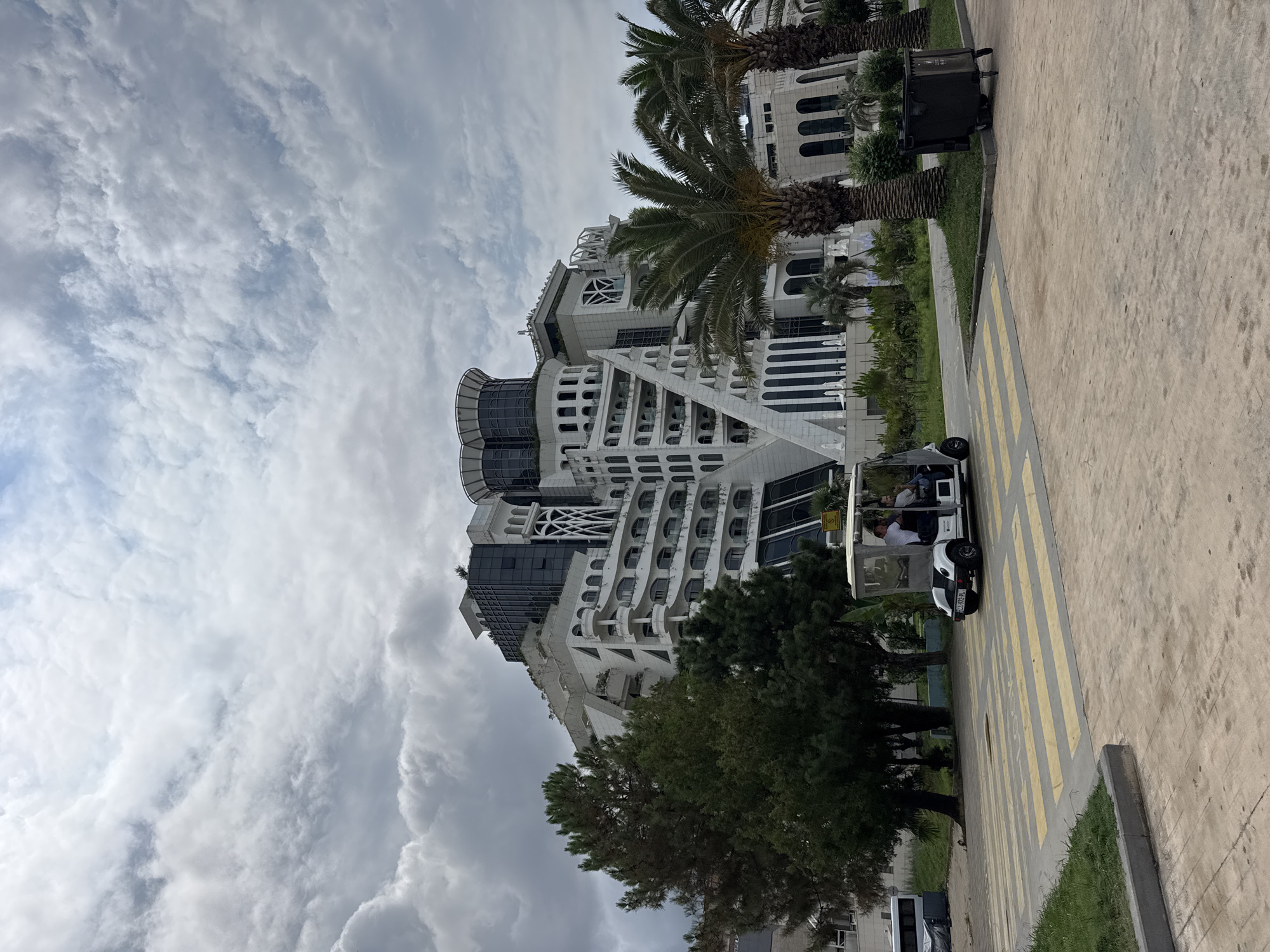 In Batumi, Georgia, a white electric golf cart with two occupants is driving across a crosswalk in front of a large, white, modern hotel with a distinctive tiered design and numerous balconies. The hotel features a unique architectural style with arched windows and decorative white columns. Two palm trees are visible to the right of the hotel, and a large dark green trash bin is situated on the grass next to them. In the background, a white bus is parked. The sky is overcast with heavy clouds, suggesting a cloudy day. The scene appears to be a resort or hotel complex, with the golf cart likely used for transporting guests around the property.