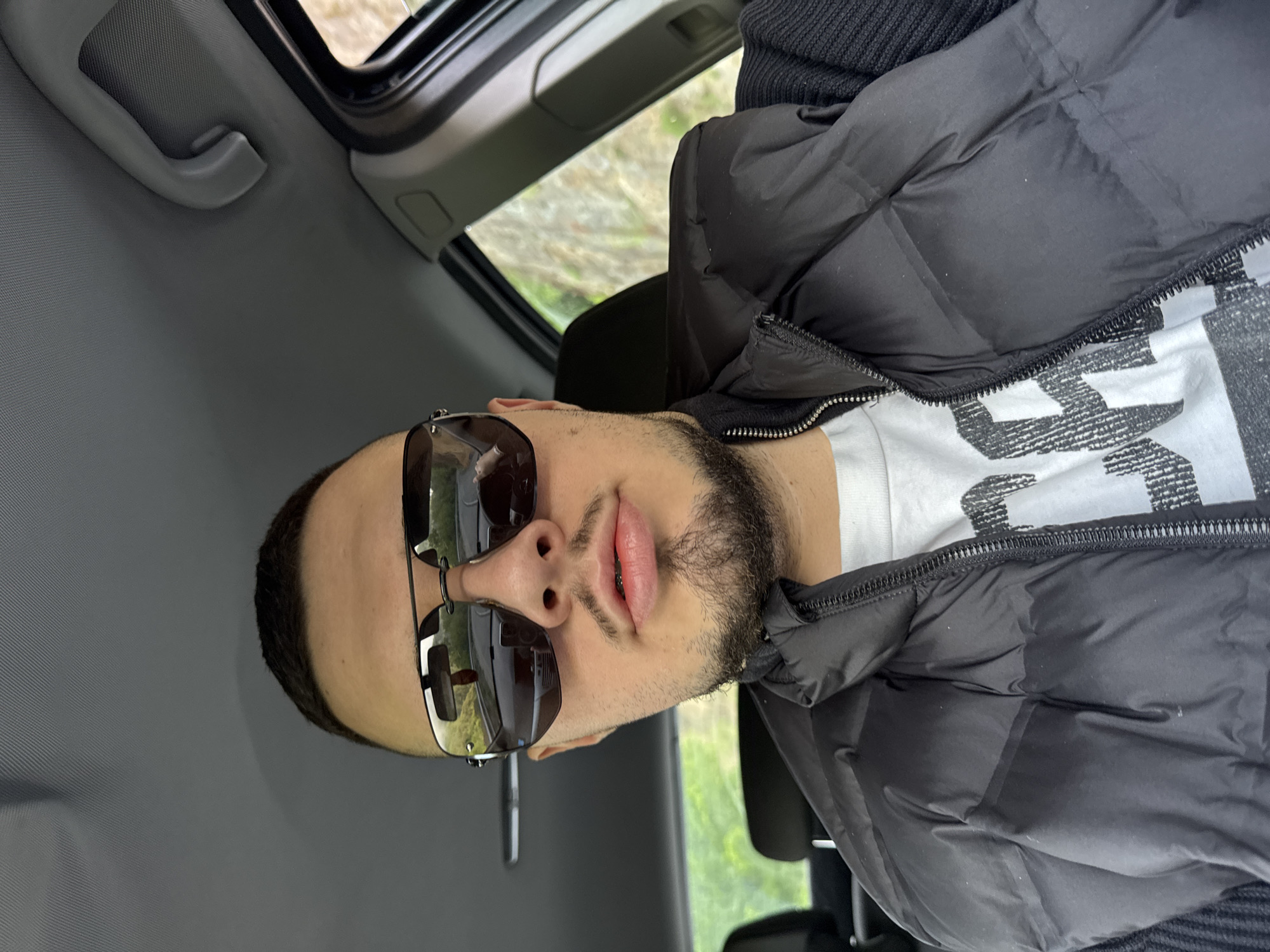 A man is photographed inside a car. He wears sunglasses, a black puffer jacket, and a white shirt. The interior of the car is gray. Outside the window, a blurry view of trees and greenery is visible. The location in the city of Taran, Bulgaria is not apparent in the image.