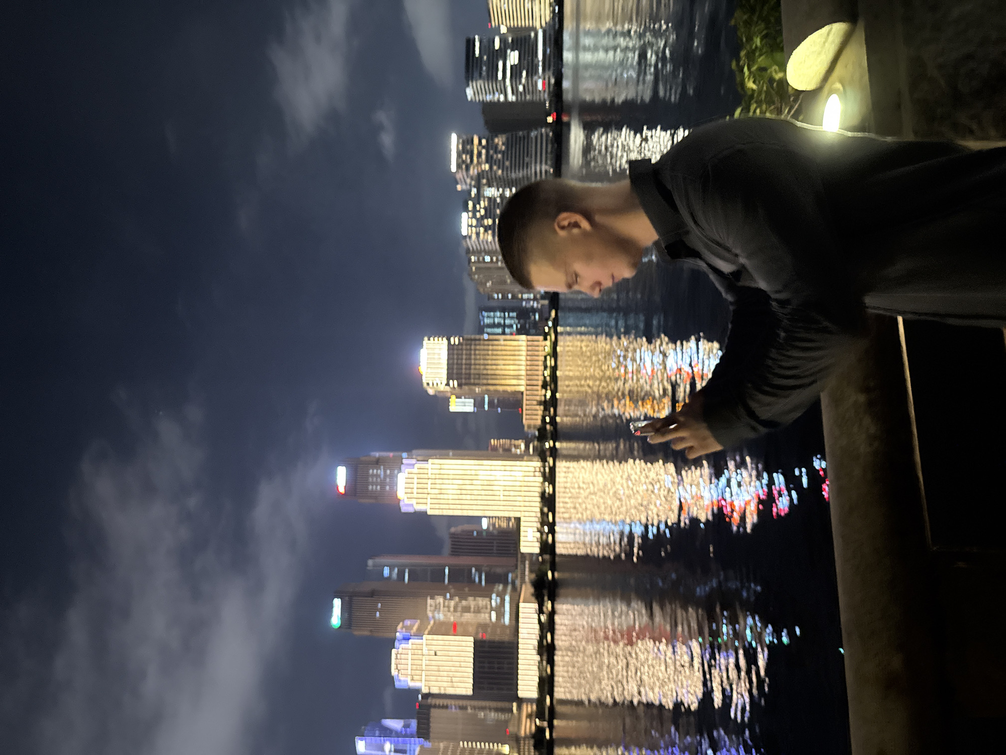 The image depicts a nighttime scene in Haizhu District, China, showcasing a modern cityscape across a body of water. In the foreground on the right, a light-skinned male with a short, faded haircut is observed in profile, facing left. He wears a dark, long-sleeved collared shirt and holds a light-colored smartphone with both hands, appearing to photograph or video the illuminated skyline. His face is faintly lit by the phone's screen, and his back is illuminated by a warm light source positioned to his immediate right on a dark, textured wall or railing. The background features numerous tall, modern buildings brightly lit with various colors, predominantly warm white, green, and red. These building lights cast distinct, shimmering reflections on the dark water surface, which occupies the midground. The sky above is dark blue-grey with visible cloud formations.