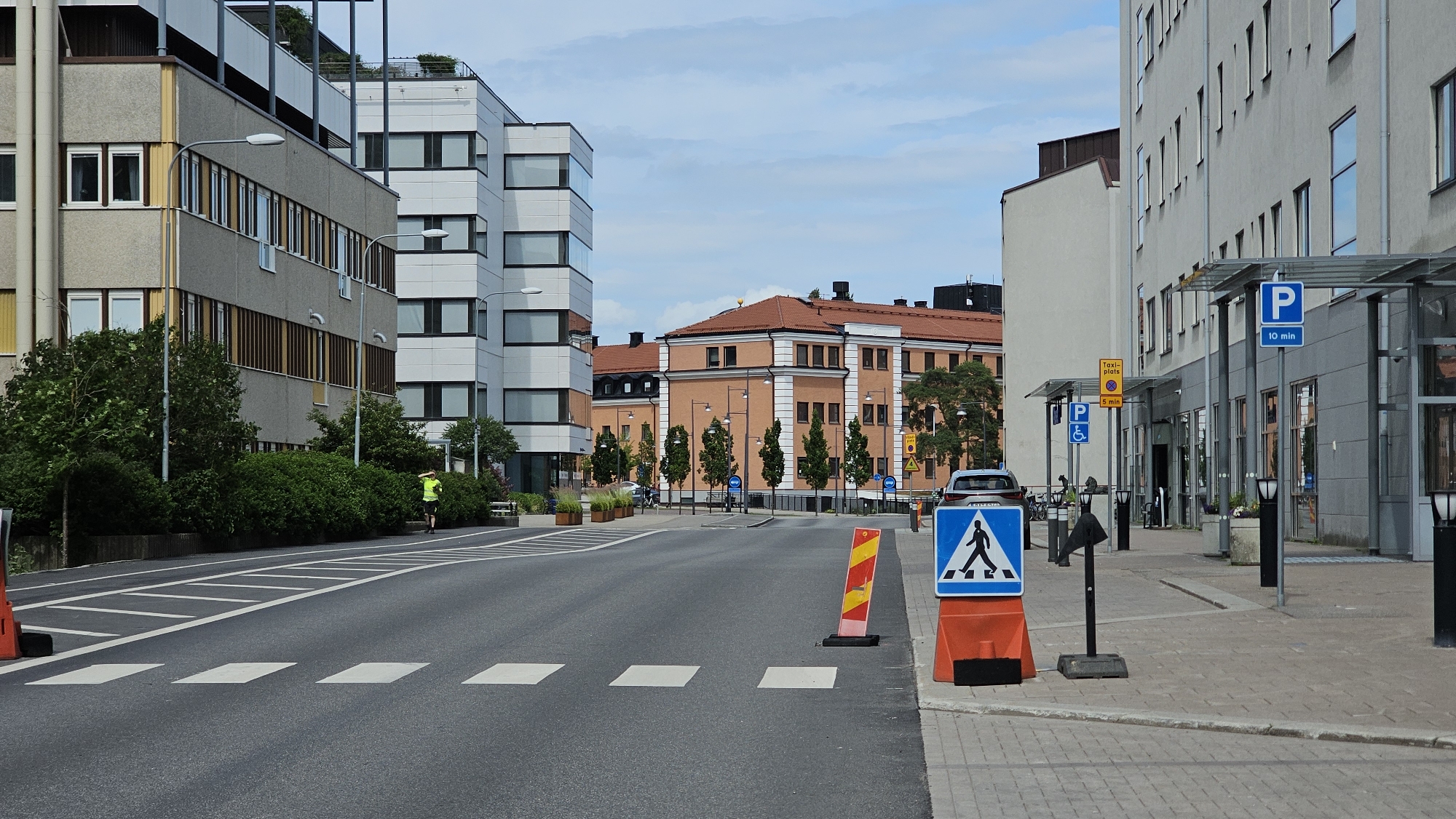 The image depicts a street scene in Linköping, Sweden, likely near the travel center. On the left, a modern grey and beige building with multiple floors and prominent windows lines the street. Adjacent to it, a taller, white building with a distinct architectural style stands. The street itself is paved and marked with pedestrian crossings. A lone individual in a bright green shirt is jogging on the sidewalk on the left side of the road.
In the background, an older, multi-story building with a terracotta-colored roof and white architectural details is visible. This building features numerous windows and appears to be part of an urban complex. Several trees are planted along the sidewalk in front of this building.
On the right side of the street, a grey building with a modern canopy and large windows dominates the foreground. A blue parking sign with a "P" and "10 min" is affixed to a pillar under the canopy. Further down the sidewalk, a yellow sign indicating "Taxi-plats" with "5 min" is visible. A blue accessibility sign is also present.
Several traffic and informational signs are strategically placed along the sidewalk. A prominent blue pedestrian crossing sign, mounted on an orange barrier, is positioned on the right side of the street, facing oncoming traffic. Another striped orange and yellow barrier is located on the road near the pedestrian crossing.
The sky is partly cloudy. The overall impression is one of a well-maintained urban environment with a mix of modern and older architecture, suggesting a functional area within the city.