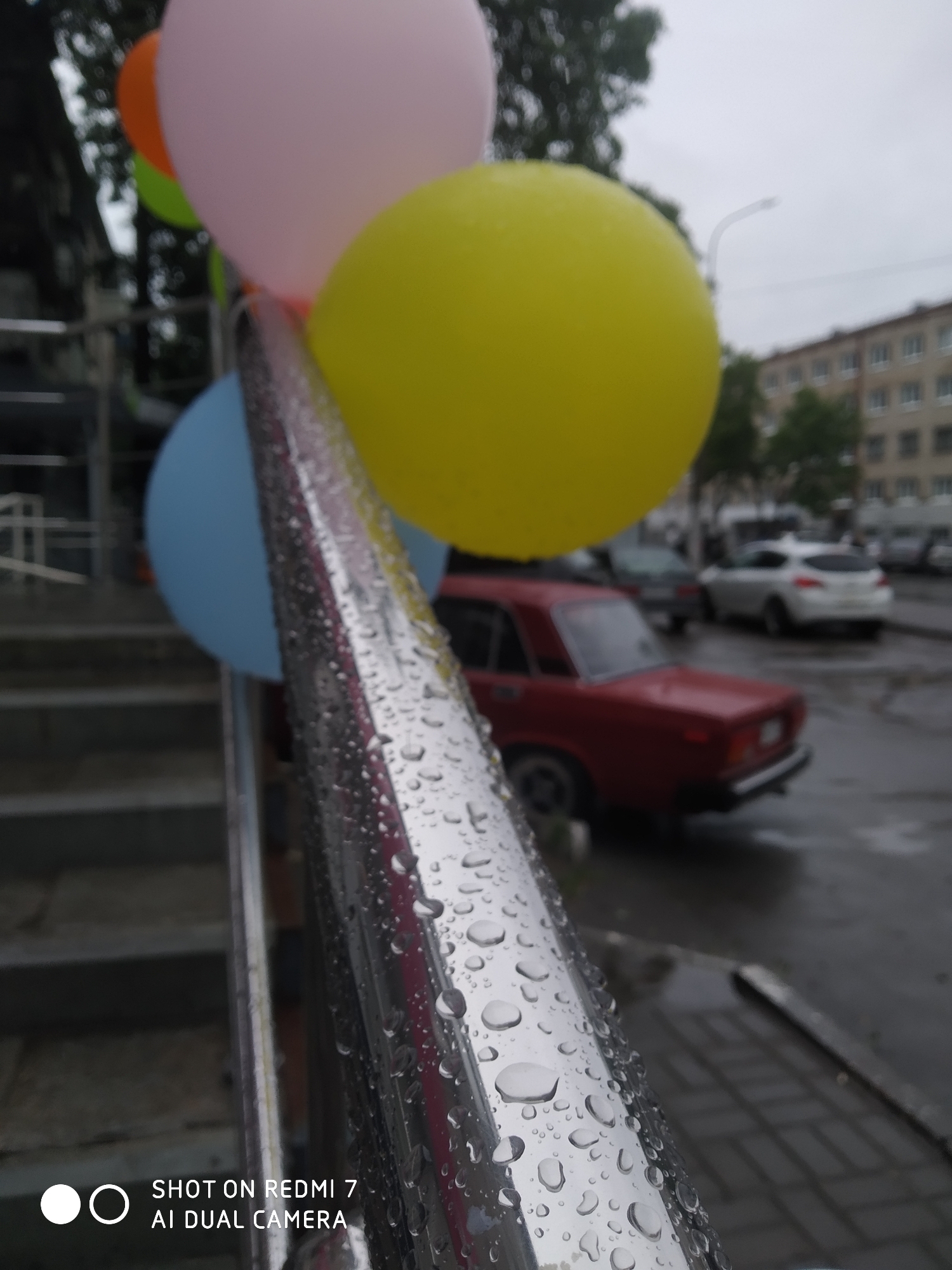 The image, taken with a Redmi 7 AI Dual Camera, depicts a rainy scene in what appears to be the city of Chaikovsky, Russia. The foreground features a chrome railing covered in raindrops, with a stack of colorful balloons tied to it (pink, yellow, blue, orange, and green visible). The background is blurred but shows a street scene with a red car, other parked vehicles, and an apartment building typical of Russian architecture. The weather seems overcast. The camera's perspective focuses on the details of the railing and balloons while indicating a residential or commercial area.