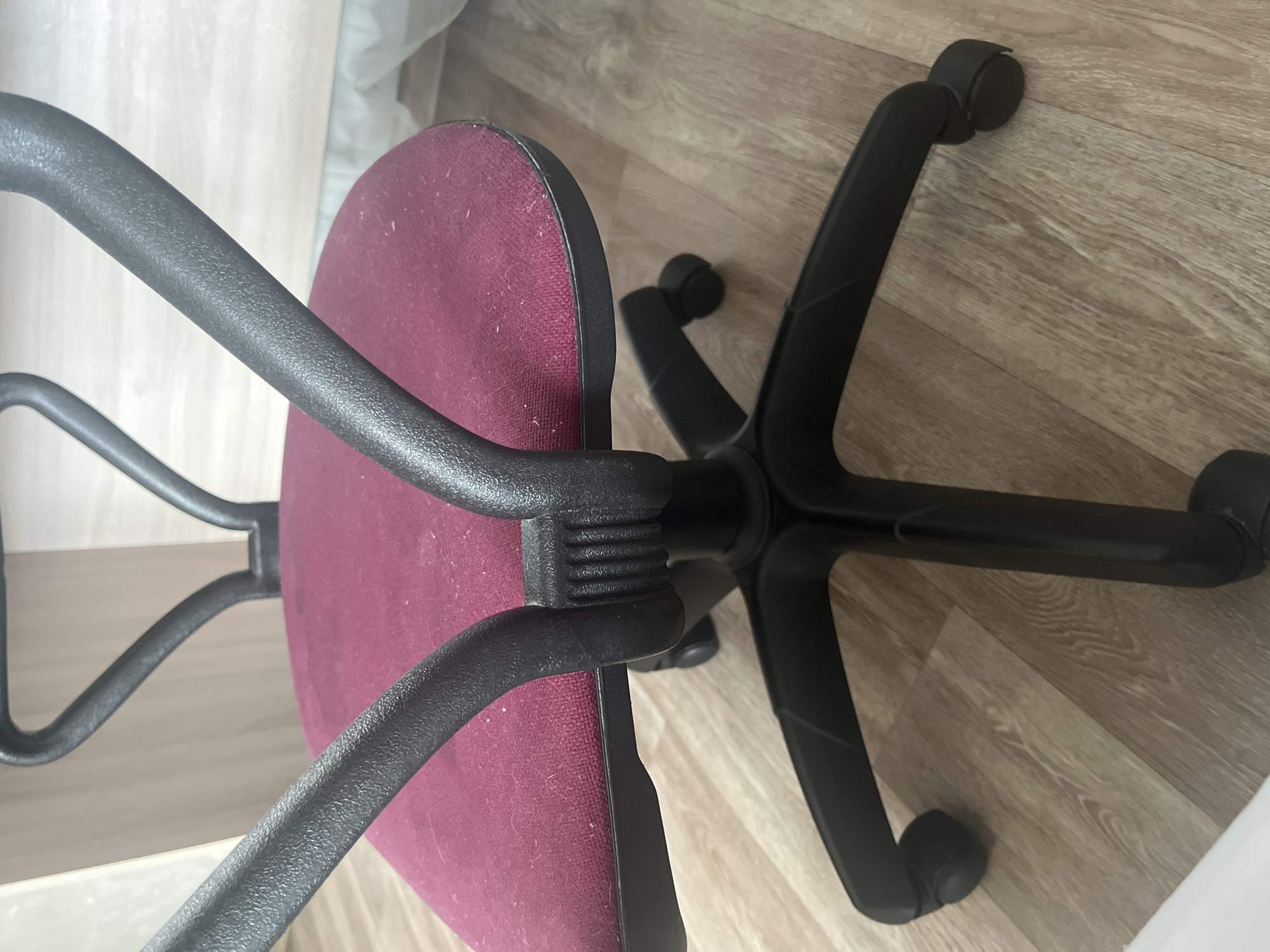 The image shows a close-up of a maroon office chair with black plastic armrests and a black five-star base with wheels. The chair is situated on a light-colored wooden floor. The scene is devoid of people, action, or interaction. The environment appears to be indoors, possibly an office or study. The exact location in Novocheboksarsk, Russia, cannot be determined from the image alone.