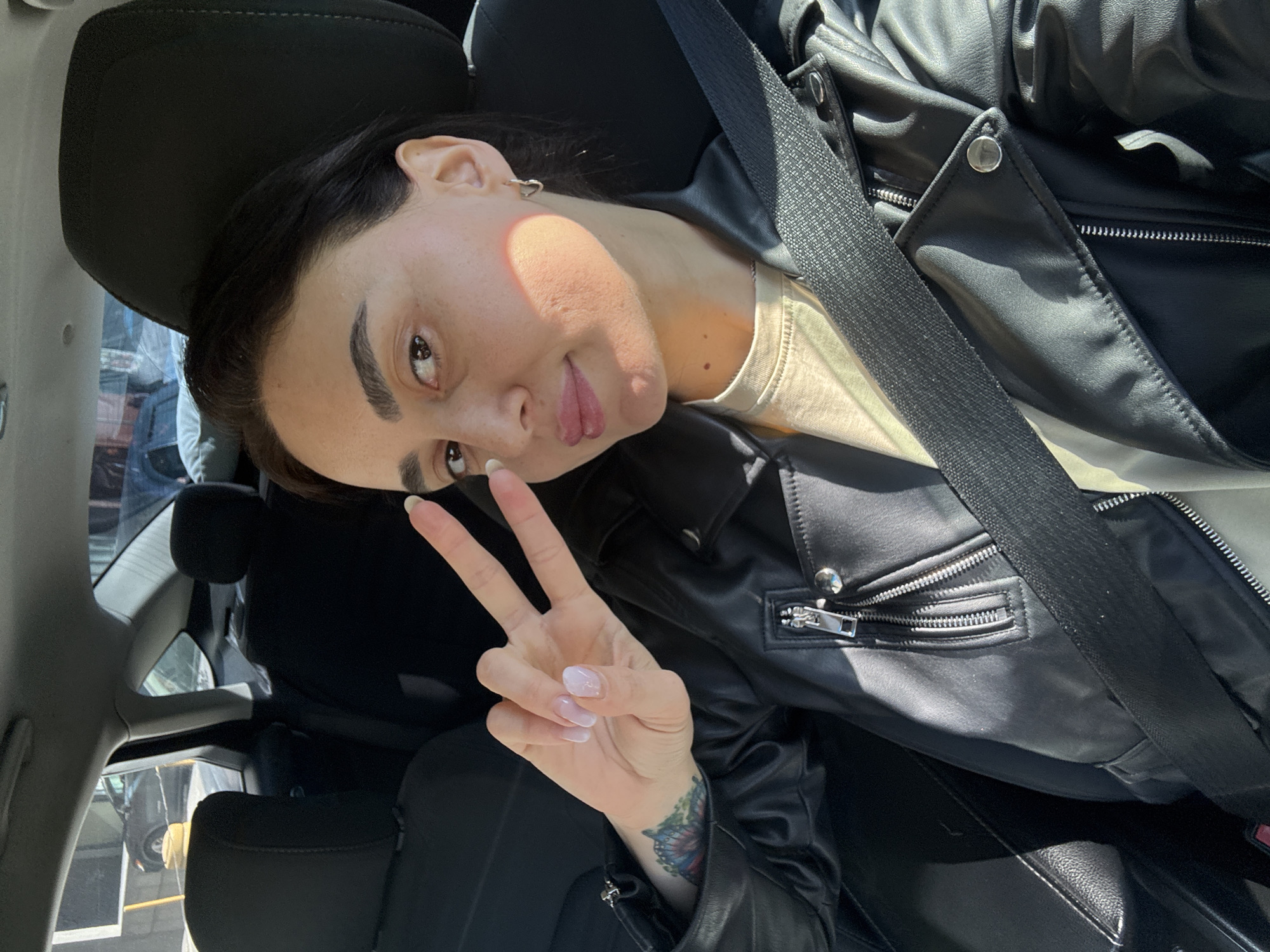 A woman is in a car in Tbilisi, Georgia, wearing a black leather jacket and seatbelt. She is holding up a peace sign. She has dark hair, fair skin, and a tattoo on her wrist. The car has black leather seats. The time of day appears to be daytime.