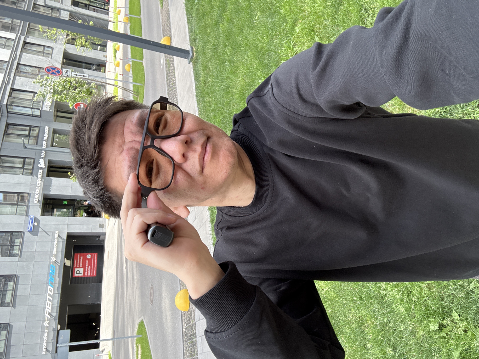 In a daylight outdoor selfie, a man with dark hair and glasses, wearing a black sweatshirt, holds up a small black object near his right eye, adjusting his glasses. He is standing in front of a modern building with a gray facade, a sign that reads "автомойка АВТОДЕТАЙЛИНГ шиномонтаж" (car wash, car detailing, tire fitting) is visible. Further to the right, signs for "ЖЕНСКИЕ СТРИЖКИ" (women's haircuts), "Soda", "MAN", "МАЙОР ПЕДИКЮР" (Major Pedicure) indicate businesses. A red parking sign with Russian text and a "P" symbol is displayed on a building entrance. The background includes a paved walkway, green grass, yellow bollards, and trees. The overall scene suggests an urban environment in Moscow, Russia.