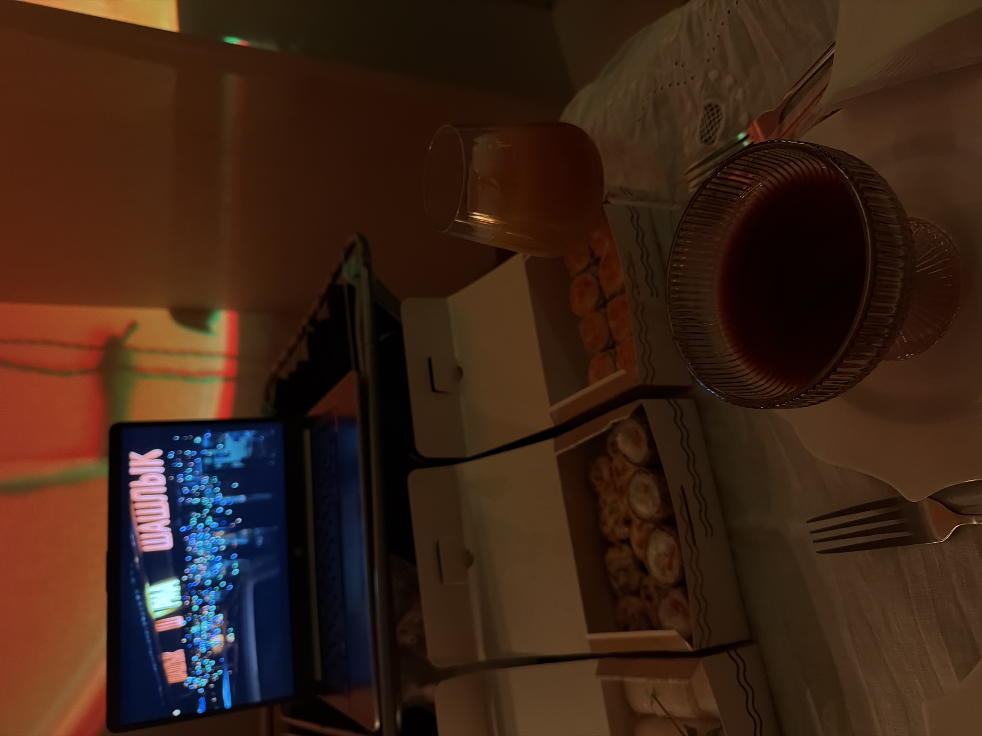 The image depicts a dimly lit indoor scene. A laptop displays the word "ШАШЛЫК" (shashlyk) in what appears to be a cityscape at night. Adjacent to the laptop are several cardboard boxes filled with sushi or similar food items. On a white tablecloth, there's a glass of amber-colored liquid and a glass bowl filled with a dark red liquid, along with silverware. Based on the text on the laptop, the scene might relate to the city of Izhevsk, Russia, given "шашлык" (shashlyk) is a popular type of food in Russia. The overall setting suggests a meal being enjoyed while watching content on the laptop.