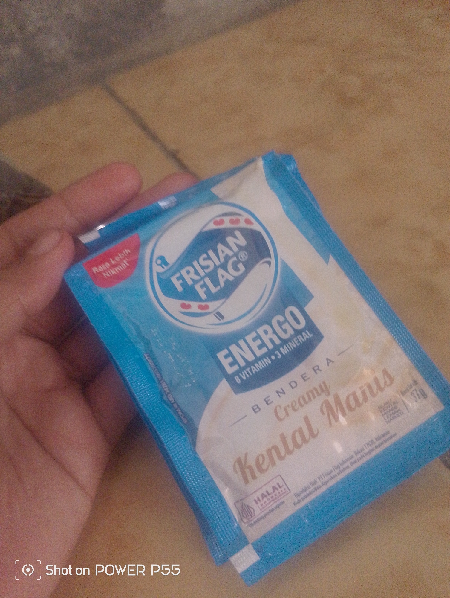 A hand is holding a sachet of Frisian Flag Energo brand creamy milk powder. The sachet is blue and white, with text in Indonesian. The background is a tiled floor. There is no indication of the city of Lebaksiu in the image.