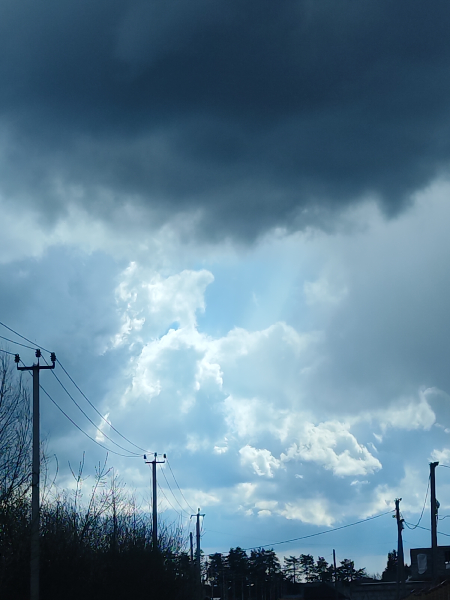 The image captures an outdoor scene dominated by a dramatic sky filled with contrasting clouds, likely in Chaikovsky, Russia. The upper and left portions of the frame are covered by dark, heavy storm clouds, suggesting overcast or tumultuous weather. Below these, and extending across the middle and right sections, are brighter, voluminous cumulus clouds, illuminated by what appears to be sunlight breaking through, revealing patches of lighter blue sky. This interplay of light and shadow creates a dynamic and somewhat foreboding yet beautiful atmosphere.
In the lower part of the frame, several dark silhouettes of utility poles with multiple overhead power lines are visible, extending across the foreground and mid-ground. Dark, indistinct silhouettes of bare tree branches or bushes are visible around the base of the poles and in the lower-left corner. Further along the horizon, especially on the right side, there are dark silhouettes of evergreen trees and some unidentifiable building structures.
The time of day appears to be daytime, characterized by the light filtering through the clouds. No human activity or visible text is present in the image. The scene combines natural elements with infrastructure, hinting at a rural or semi-urban setting under changing weather conditions.