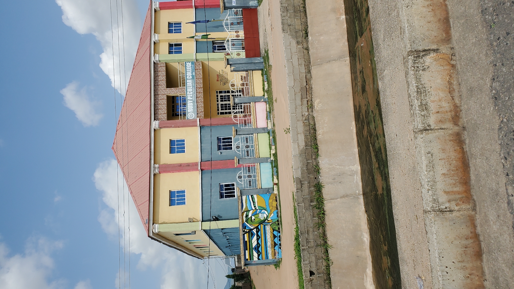 The image shows the exterior of Ruby Peculiar College in Jalingo, Nigeria. The building is a multi-story structure painted in yellow, blue, and red. It has a red-tiled roof and several windows on each floor. In front of the building is a fence with colorful sections and decorative metalwork. To the left of the building, a mural depicting a person's face and abstract patterns is visible on a wall. A concrete drainage channel runs in front of the fence, with murky water and green algae present. The sky above is blue with scattered white clouds. Power lines are visible against the sky.