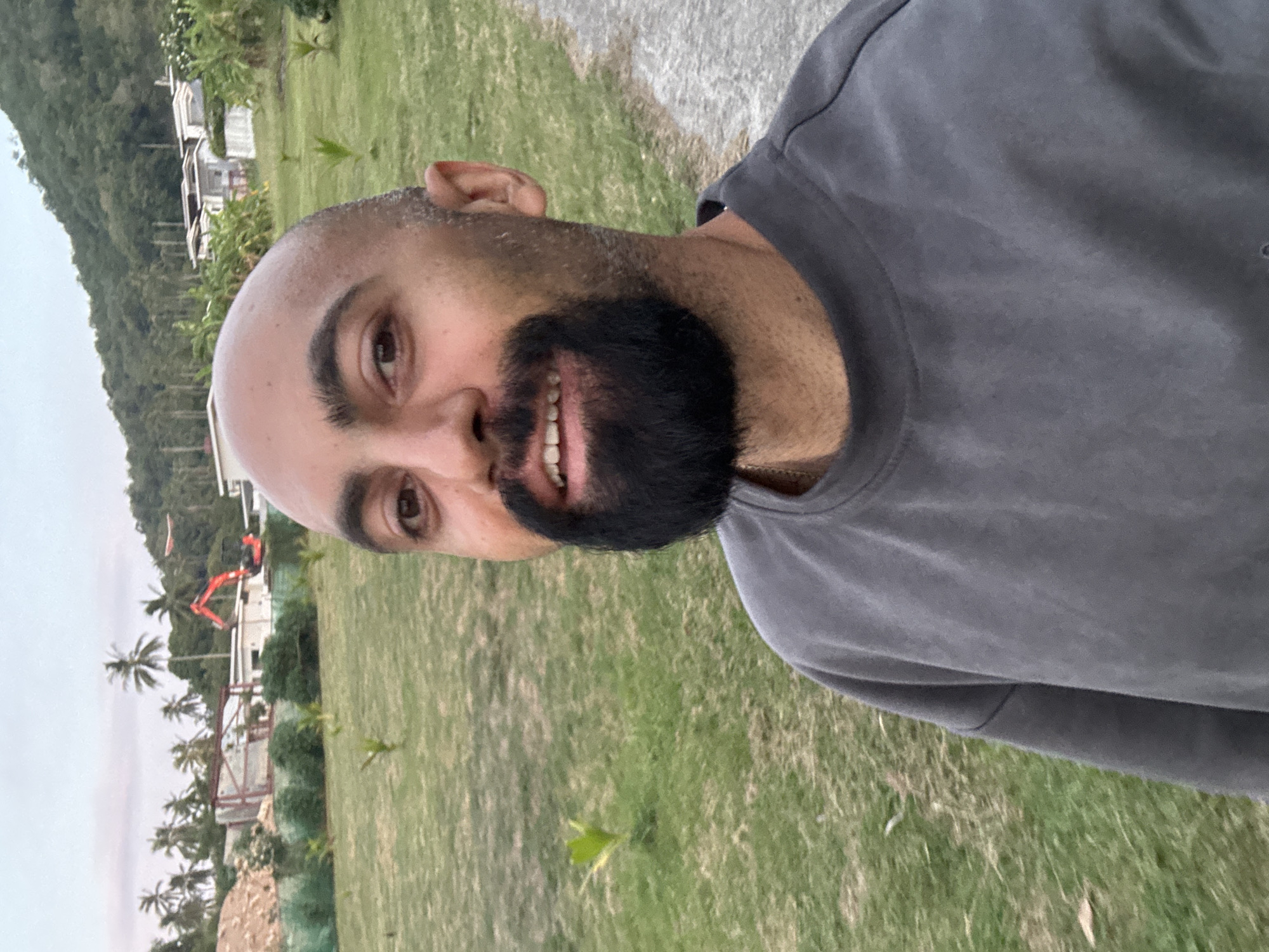 The image is a selfie of a bald man with a beard, wearing a dark gray sweatshirt. He is standing outdoors in front of a grassy area with scattered young palm trees. In the background, there is a construction site with an excavator, piles of dirt, and unfinished buildings. Further in the distance, there are palm trees, houses, and a lush green hillside. The sky is overcast, suggesting it might be early morning or late afternoon. The scene appears to be in a tropical location.