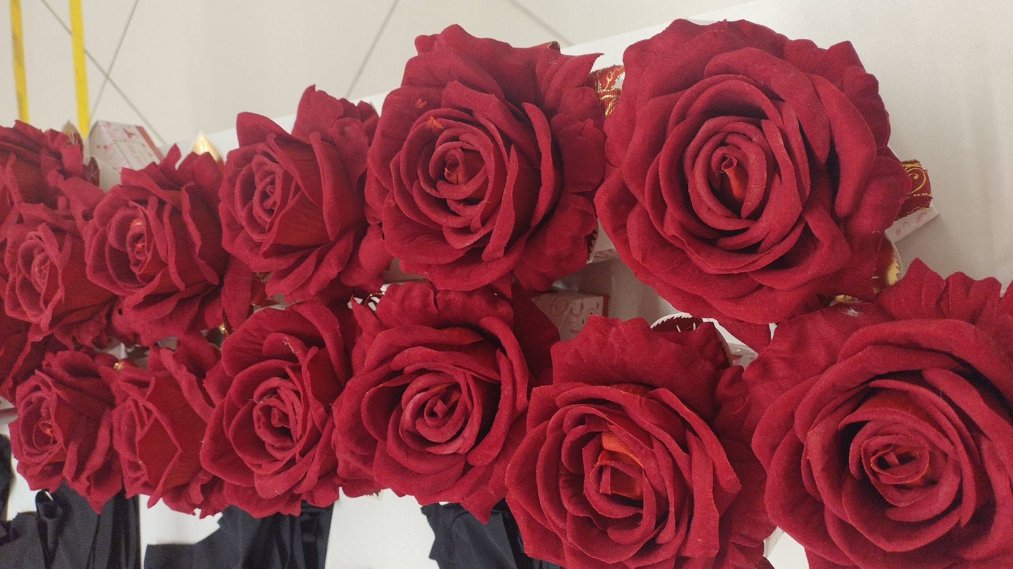 A top-down, eye-level shot shows a display of artificial red roses arranged in multiple rows. The roses are a deep, velvety red with intricate petal formations, and some appear to have gold trim at the base. The background is a white surface, likely a table, with hints of black fabric on the left side. A yellow line can be seen in the distance, suggesting a marked area on a floor or wall. The setting appears to be indoors, possibly a retail or display area. The roses are the primary focus, presented in a visually rich and orderly fashion.