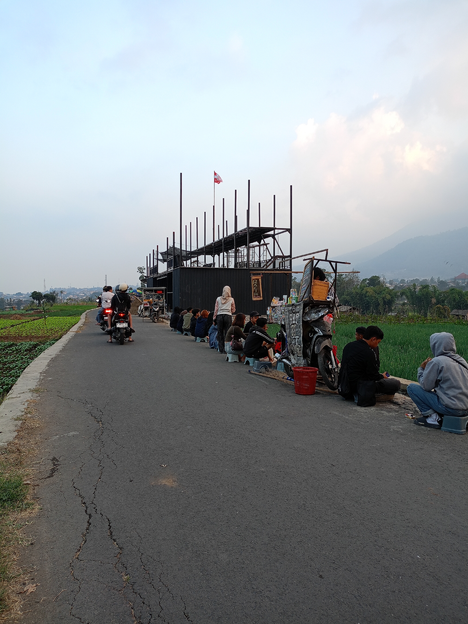 In Batu, Indonesia, a group of individuals are gathered along the side of a cracked asphalt road. To the right, a modern, multi-level structure with a black facade and a dark metal frame stands, topped with a small flag. In front of this building, several people are seated on small blue stools, facing the road. A motorcycle with a cart attached is parked nearby. Further down the road, another group of people are riding motorcycles. To the left of the road, lush green fields stretch out, bordered by a concrete edge. In the background, a misty mountain range overlooks the scene under a pale, cloudy sky.