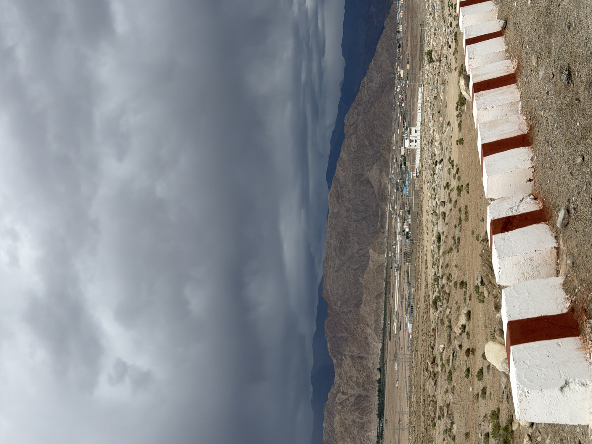 The image depicts a mountainous landscape under a cloudy sky. In the foreground, a series of concrete barriers painted with red and white stripes line a road. The barriers are weathered and appear to be part of road safety infrastructure. Beyond the barriers, a dry, rocky terrain stretches towards a town situated at the foot of large, brown mountains. The town appears to have a mix of residential and commercial buildings, with some trees and vegetation visible. In the background, several layers of mountains recede into the distance, their peaks obscured by the heavy, dark clouds that dominate the sky, suggesting an impending storm or overcast weather conditions. There are no people or animals visible in the image. The overall scene conveys a sense of a remote, arid environment with a hint of approaching weather change.