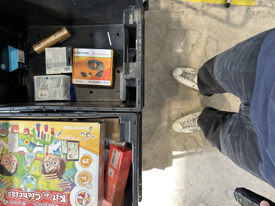 This top-down image depicts a person standing on a concrete floor in the Polígon Industrial De Son Castello, Spain, looking down into two open, black plastic storage crates positioned directly in front of them. The person is wearing a dark blue shirt, dark grey wide-leg pants, and off-white sneakers with a star emblem. They are holding a black-cased smartphone in their left hand. The concrete floor shows wear, cracks, and some irregular discoloration, with a yellow marking line visible on the right.
The left crate contains a large, colorful box for a "Kit de Ciencias" (Science Kit) with cartoon illustrations of children, positioned atop a smaller red rectangular box. The right crate contains an assortment of items, including an HP 303 ink cartridge, a gold-colored aerosol can of L'Oreal hair product, several small packaged items (likely electronic components), and an orange and black box labeled "GR.FEMA Bike Cable Lock." A small white slip of paper is also visible within the right crate. Strong overhead lighting casts shadows of the crates and the person onto the floor.
