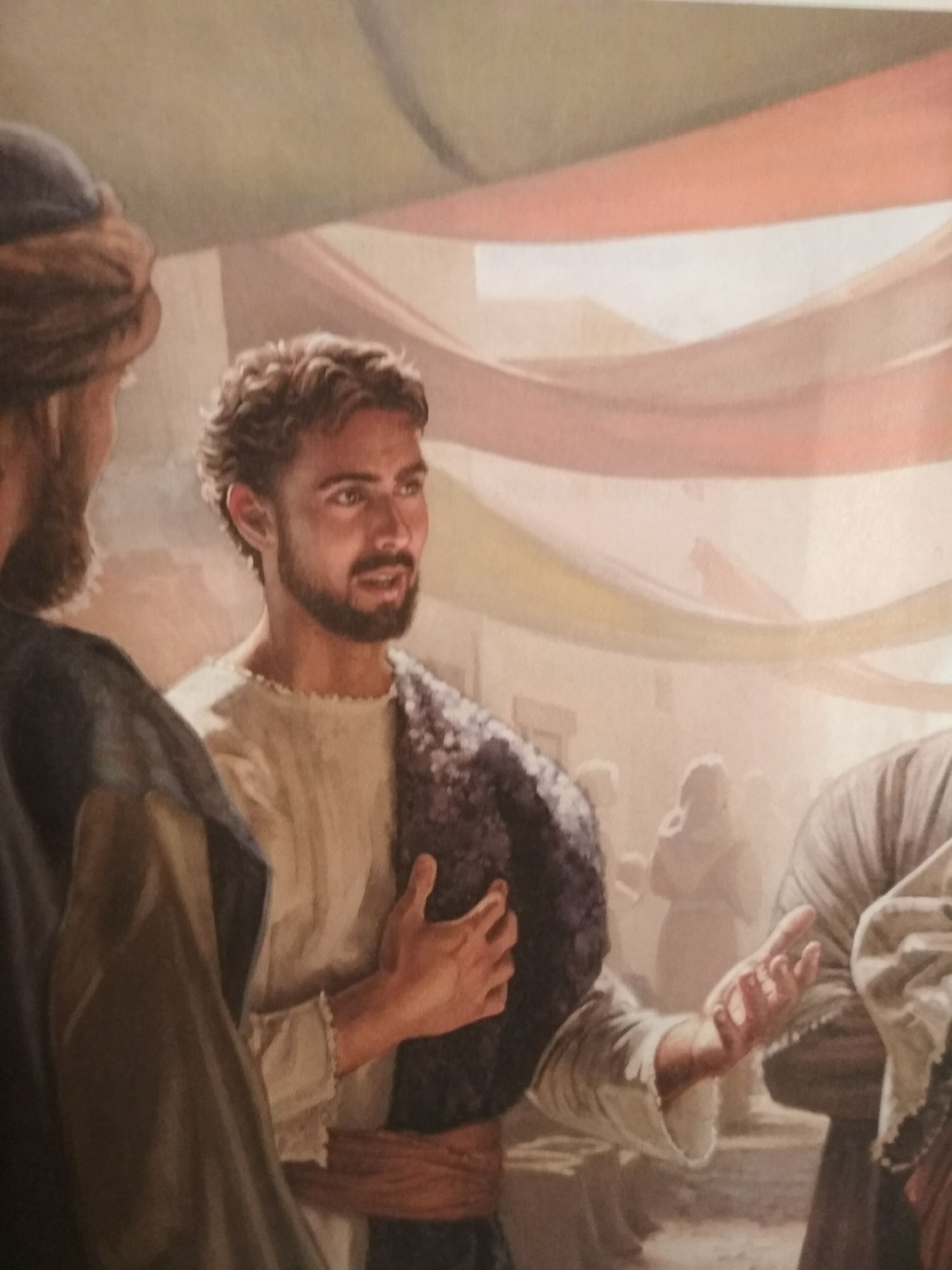 The image depicts a painting of a man with curly brown hair, a beard, and dark eyes, dressed in a light-colored tunic with a dark sash. He has a dark shawl draped over one shoulder. He is gesturing with his right hand as if speaking or explaining something. He is surrounded by other people, with parts of them visible on either side of him. The setting appears to be outdoors, with colorful banners or decorations hanging above. The architecture and the presence of draped cloth suggest a market setting. The painting could be set in the city of Gura Top, Nigeria.
