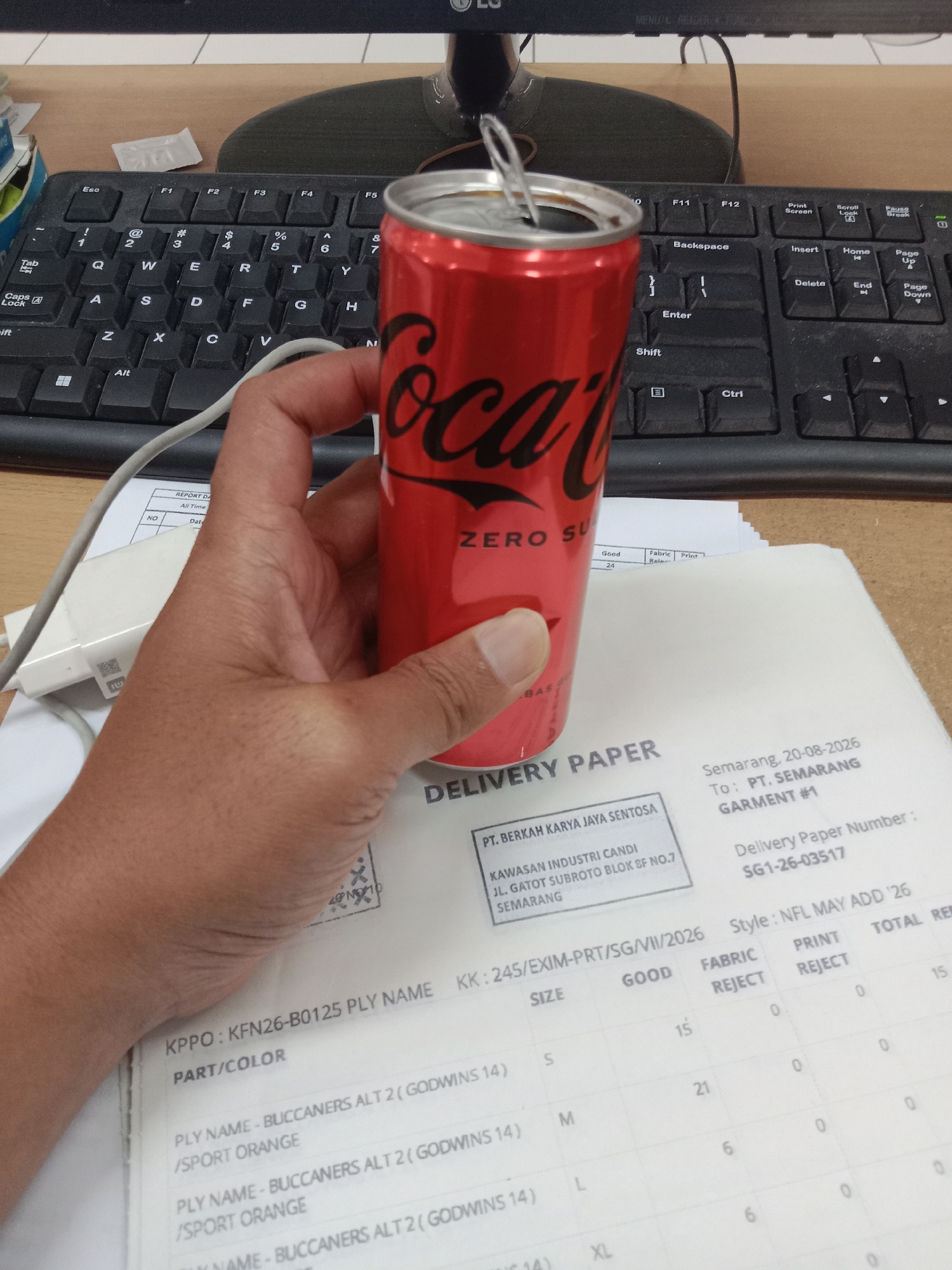 The image shows a hand holding a Coca-Cola Zero Sugar can over a delivery paper. The delivery paper has "DELIVERY PAPER" printed at the top. Below that, there is a section with "PT. BERKAH KARYA JAYA SENTOSA" and an address that includes "KAWASAN INDUSTRI CANDI JL. GATOT SUBROTO BLOK 8F NO.7 SEMARANG". To the right of this section, there is a header indicating "Semarang, 20-08-2026" and "To: PT. SEMARANG GARMENT #1". The paper also lists "Delivery Paper Number: SG1-26-03517" and a table with columns for "SIZE", "GOOD", "FABRIC REJECT", "PRINT REJECT", and "TOTAL REJECT". The hand is positioned to obscure some of the details on the paper, but some entries are visible, such as "S" under SIZE with "15" under GOOD and "XL" under SIZE with "6" under GOOD. The keyboard of a computer is visible in the background, with the Coca-Cola can and hand in the foreground. The environment appears to be an office or workspace.