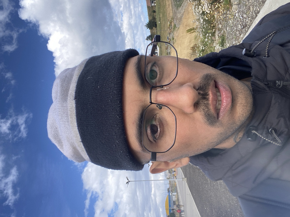 A man wearing glasses and a striped beanie poses for a selfie. He is also wearing a dark colored jacket. In the background, a cloudy blue sky is visible on the left, contrasting with a beige field dotted with small white flowers on the right. A road with lampposts is seen beneath him, adding depth to the outdoor scene.