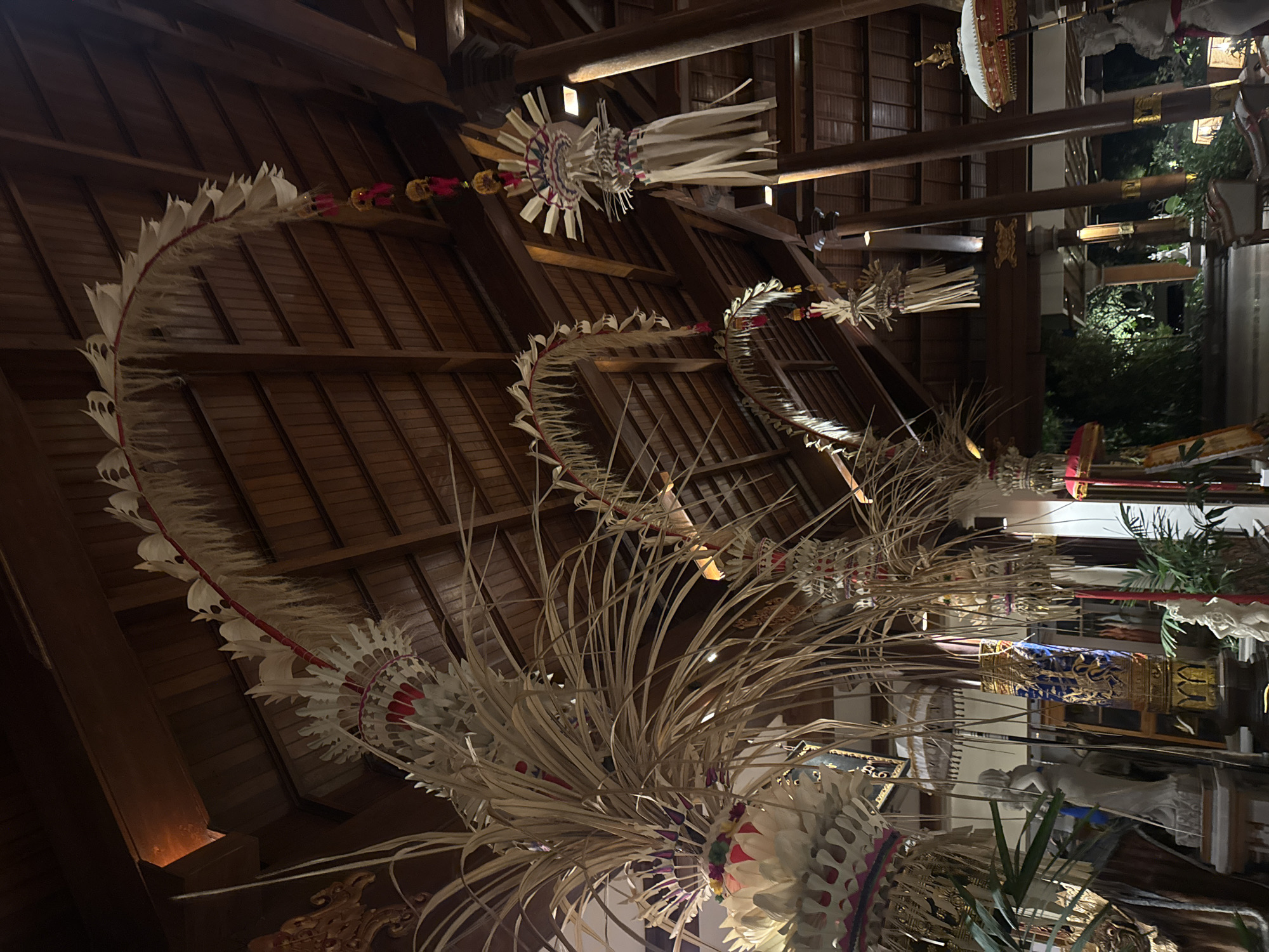 The image shows a dimly lit interior space with a high, wooden-beamed ceiling. Ornate, traditional Balinese decorations, crafted from dried palm leaves and fabric, hang from the ceiling and along curved structures. These decorations are elaborate, featuring intricate woven patterns, white fringed elements, and accents of red and pink.
Several hanging ornaments are visible, some with multiple tiers of woven elements and suspended beads. In the foreground, large, bushy arrangements of dried palm leaves are prominent.
The environment appears to be an indoor public space, possibly a lobby or a hall, with the architectural style suggesting a resort or cultural center in Bali. In the background, partially visible are statues, a decorative pillar with blue and gold detailing, plants, and hints of an outdoor area bathed in darkness with some artificial lighting. The overall ambiance is one of cultural decoration and a warm, inviting atmosphere, despite the low light.
