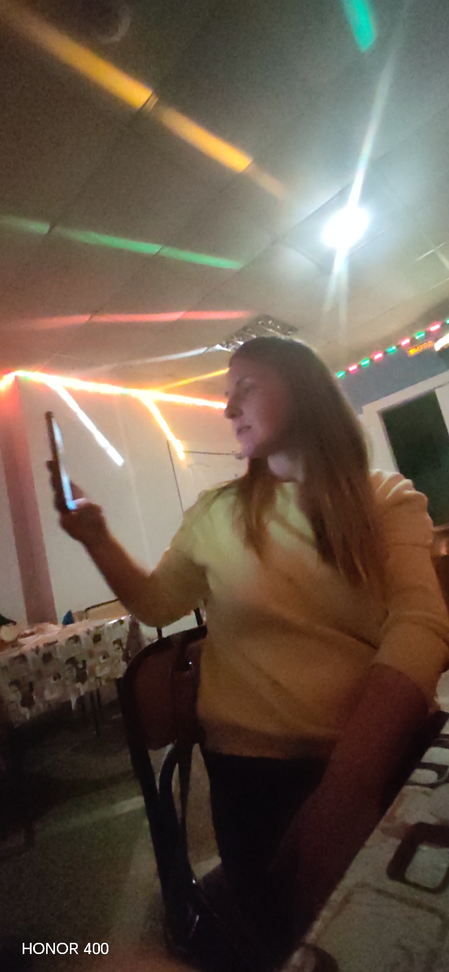 A photograph shows a woman holding a mobile phone in a room with party lighting. The woman is seated in a chair, wearing a yellow sweater. She is looking at the phone in her hand. The room is illuminated with multi-colored neon lights and a ceiling light. A table with a patterned tablecloth is visible in the background. The photo was taken with an HONOR 400 phone. The location of the photo is Noviy, Russia, as indicated by the Russian text on a sign near the window.