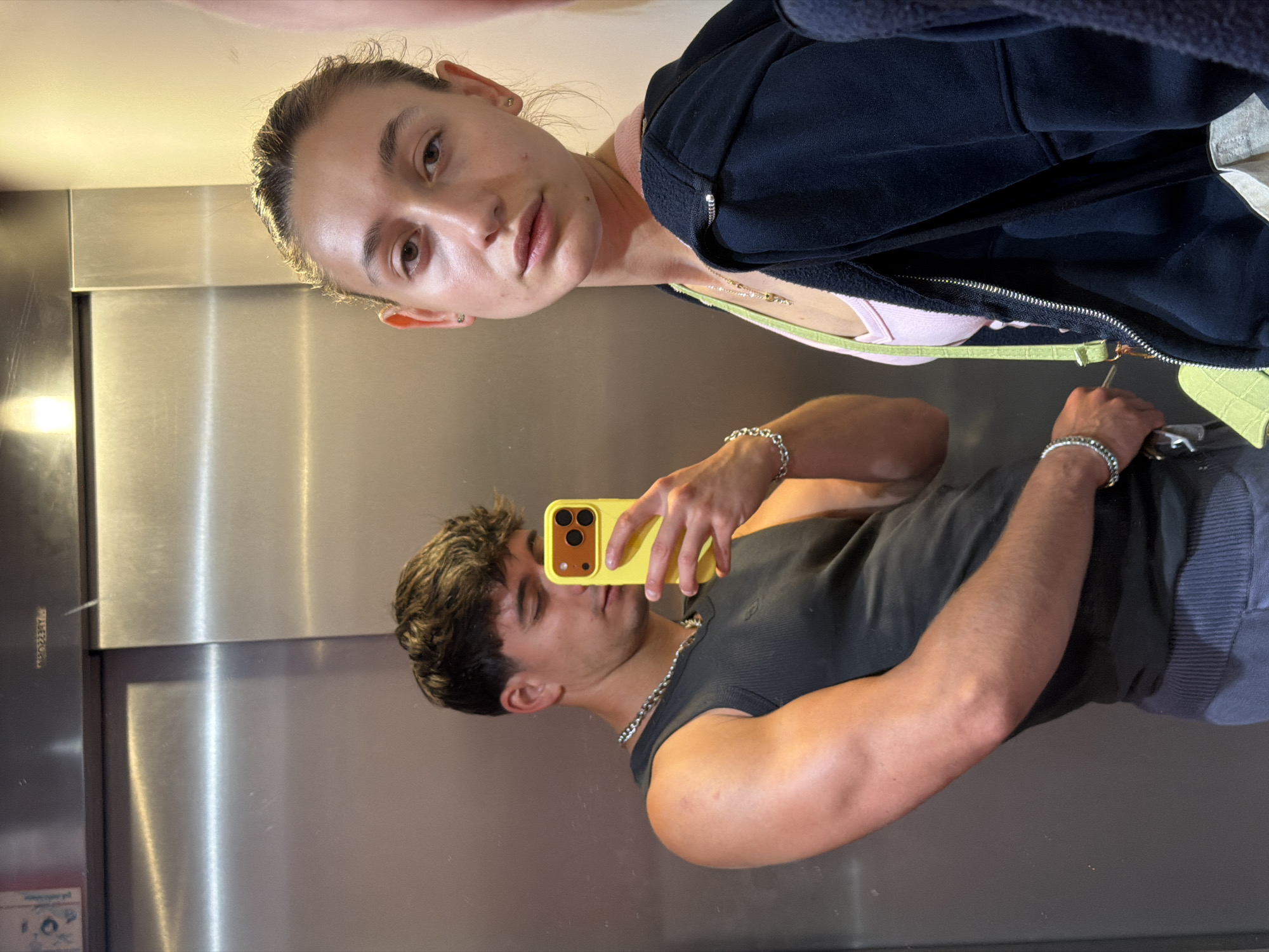 The image depicts a young man and woman in an elevator. The man, on the left, is taking a selfie with a yellow phone while looking down. He is wearing a dark sleeveless shirt and a silver chain. The woman, on the right, is looking towards the camera with a neutral expression. She is wearing a pink tank top underneath a dark zip-up hoodie and a green crossbody bag. The elevator walls are made of brushed metal, reflecting the surrounding environment. The lighting is dim and appears to be artificial. This scene does not contain any specific details that would identify its location in the city of Sofia, Bulgaria.