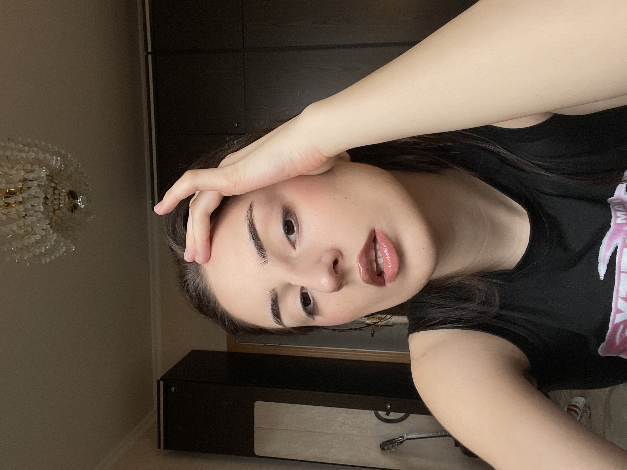 The media file presents an indoor selfie of a young woman, captured in Moscow, Russia. The image is oriented with a noticeable clockwise tilt. The woman, with dark hair and prominent dark eyebrows, is looking directly at the camera. Her right hand is placed on her forehead, gently resting on her hair. She features a subtle blush on her cheeks and wears glossy lip gloss, with her lips slightly parted to reveal some teeth. She is dressed in a black t-shirt or tank top, which has a partially visible pink and white graphic or text on the right side, possibly reading "MAL" or "MAN".
The background on the left side of the frame displays an ornate chandelier adorned with numerous clear or white crystal-like beads. The wall behind it is a light, neutral color. On the right and extending towards the bottom, dark wooden furniture is visible, appearing to be a tall wardrobe or cabinet positioned above a lower cabinet. A light-colored object, possibly a drawer or part of a suitcase, is seen beneath the lower cabinet, alongside the headstock and neck of a string instrument, resembling a guitar. A small portion of a white and red shoe is also visible on the floor on the far right. The indoor setting appears to be well-lit, suggesting either daytime or a brightly illuminated room. The woman maintains a composed and direct expression.
