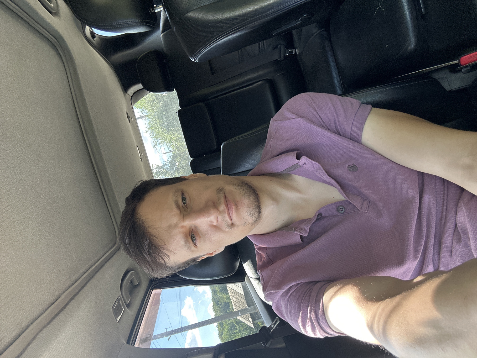 The image shows a man sitting in the back seat of a car. He is wearing a purple polo shirt and looking towards the camera. Sunlight streams in from the window, illuminating his arm and shoulder. The interior of the car is visible, including black leather seats and the car's ceiling. Through the window, a rural landscape with trees and a small house can be seen under a bright, cloudy sky, consistent with the location of Makiv, Ukraine.