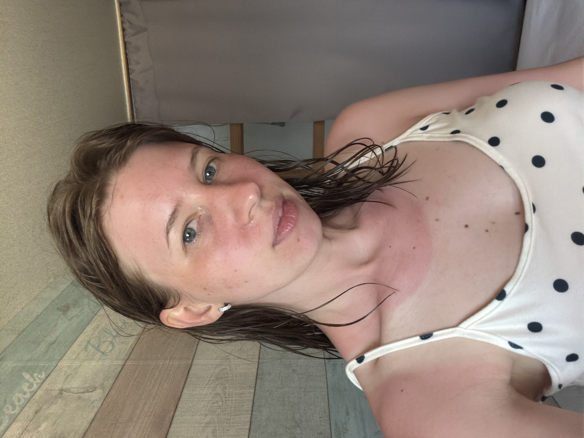 A young woman with wet brown hair and blue eyes looks directly at the camera. She is wearing a white polka dot top. Her face and neck are flushed red, indicating a sunburn. The background consists of a wooden wall with vertical planks of various colors, and a gray curtain on the right. This scene is located in Gangneung-si, South Korea.