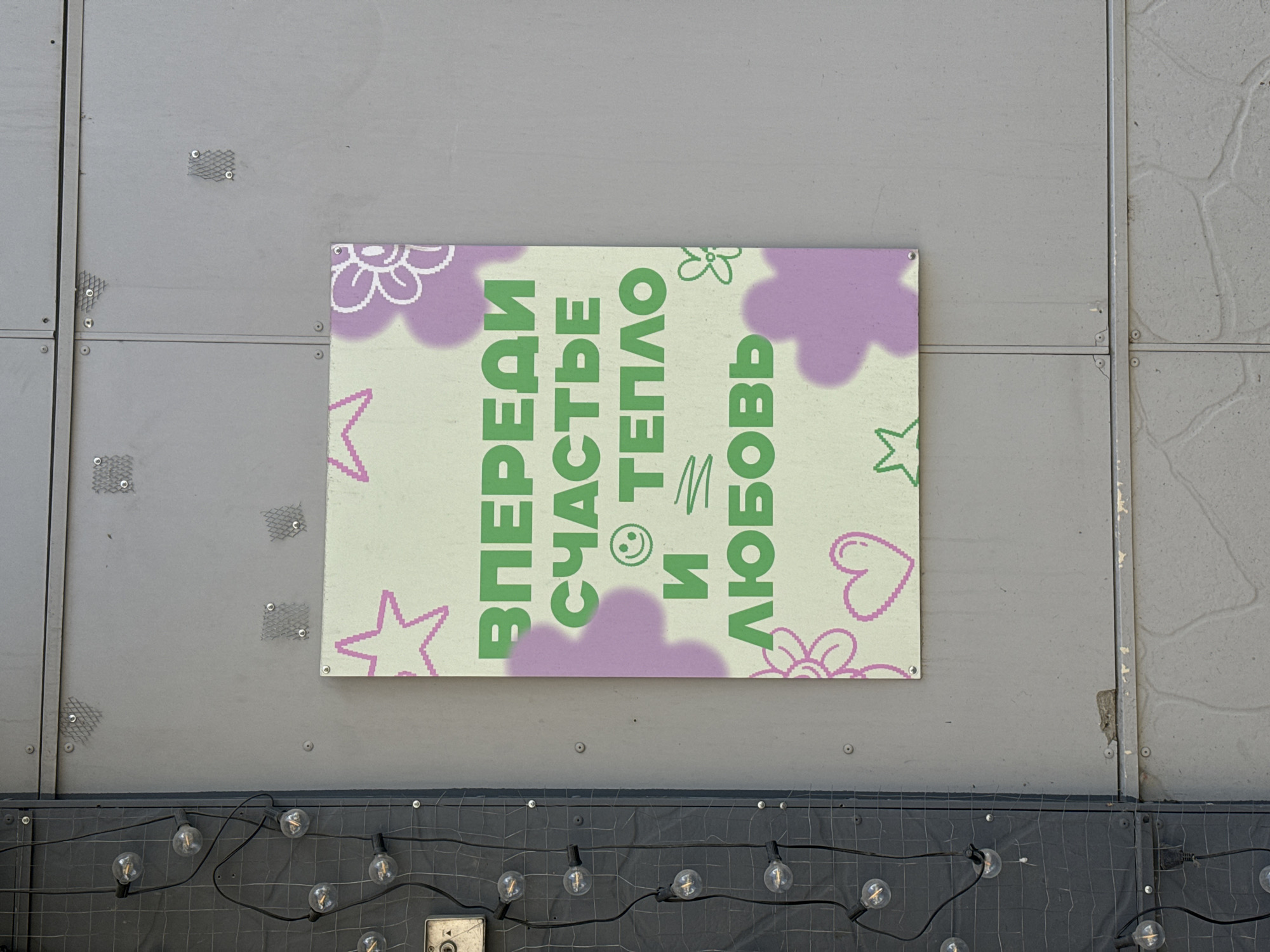 A sign hangs on a gray metal wall in what appears to be Krasnoyarsk, Russia, based on the Cyrillic text. The sign has a pale green background and features the words "ВПЕРЕДИ СЧАСТЬЕ И ТЕПЛО И ЛЮБОВЬ" in green lettering. There are purple cloud-like shapes and outlines of flowers, stars, and a heart scattered around the text. A string of light bulbs is strung across a dark surface beneath the wall. The sign is attached to the wall with screws or rivets at each corner. The metal wall shows signs of wear and has a section covered with a gray grid.