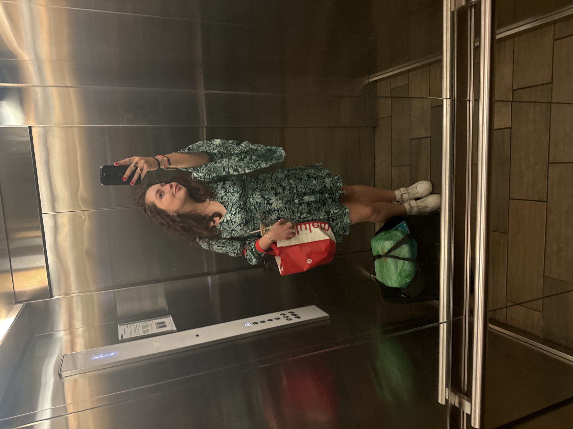 A young woman is taking a selfie in an elevator. She is wearing a green patterned dress and white sneakers. She is holding a red and white tote bag and a green bag is at her feet. The elevator has stainless steel walls and a control panel with illuminated buttons. The elevator doors are partially open, revealing a tiled floor. The location appears to be in Mexico City, Mexico, based on the "meli" on the tote bag, which likely refers to Mercado Libre, a popular e-commerce platform in Latin America.