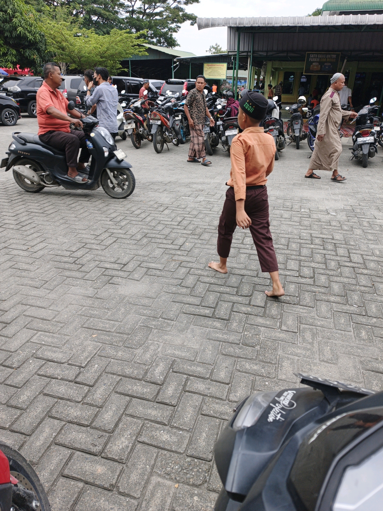 In Manunggal, Indonesia, a paved outdoor area, likely a parking lot for a community building, is depicted. The ground is covered with interlocking gray bricks laid in a herringbone pattern. Several individuals are present. In the foreground, a young boy wearing an orange shirt, dark pants, and a black cap walks barefoot. Behind him, an adult man in a light brown robe and cap walks towards the right. To the left, a man in an orange shirt and dark pants rides a black scooter. Other people are visible in the background, some standing near a building with green accents and a white roof. A row of parked motorcycles is also present, with cars further in the background. The environment suggests a sunny day, with trees visible behind the building.