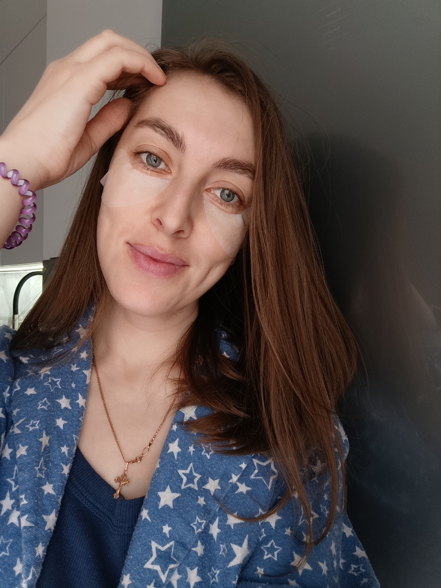 A woman with long brown hair and blue eyes is posing for a selfie. She is wearing a blue shirt with white stars and a gold necklace with a cross pendant. There are light-colored patches under her eyes. She has a purple spiral bracelet on her left wrist and is touching her forehead with her left hand. The background appears to be a dark gray surface, possibly a wall or appliance. There is not enough information available to determine the location as Chaikovsky, Russia or confirm the depicted items.