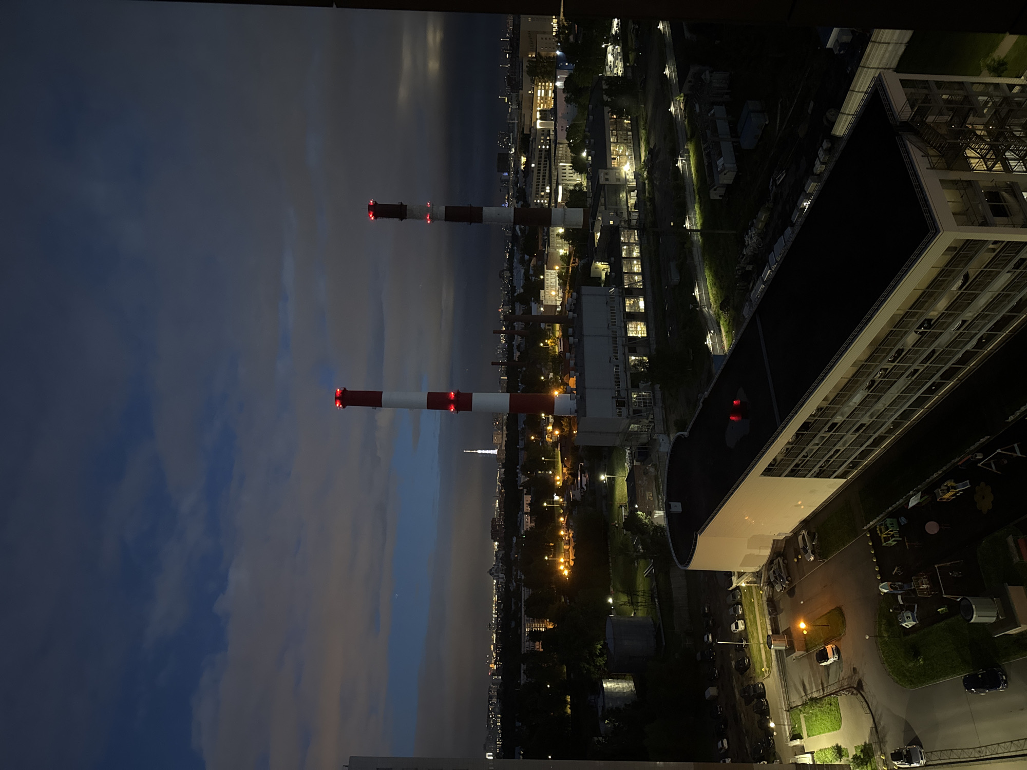 The image captures a twilight view of an industrial area in an unknown city in Russia. Dominating the scene are two tall, red and white striped smokestacks, each with red lights at the top. In the foreground, a long, white building with a dark roof is visible, with windows and an exterior fire escape. Adjacent to this building is a playground with various colorful structures. Several cars are parked in a lot nearby. Further in the background, a sprawling cityscape is illuminated by numerous lights, with a prominent tall, thin tower visible on the horizon. The sky is filled with dark, cloudy formations interspersed with patches of lighter sky, suggesting the late hours of the day. The overall impression is an urban industrial landscape under a darkening sky.