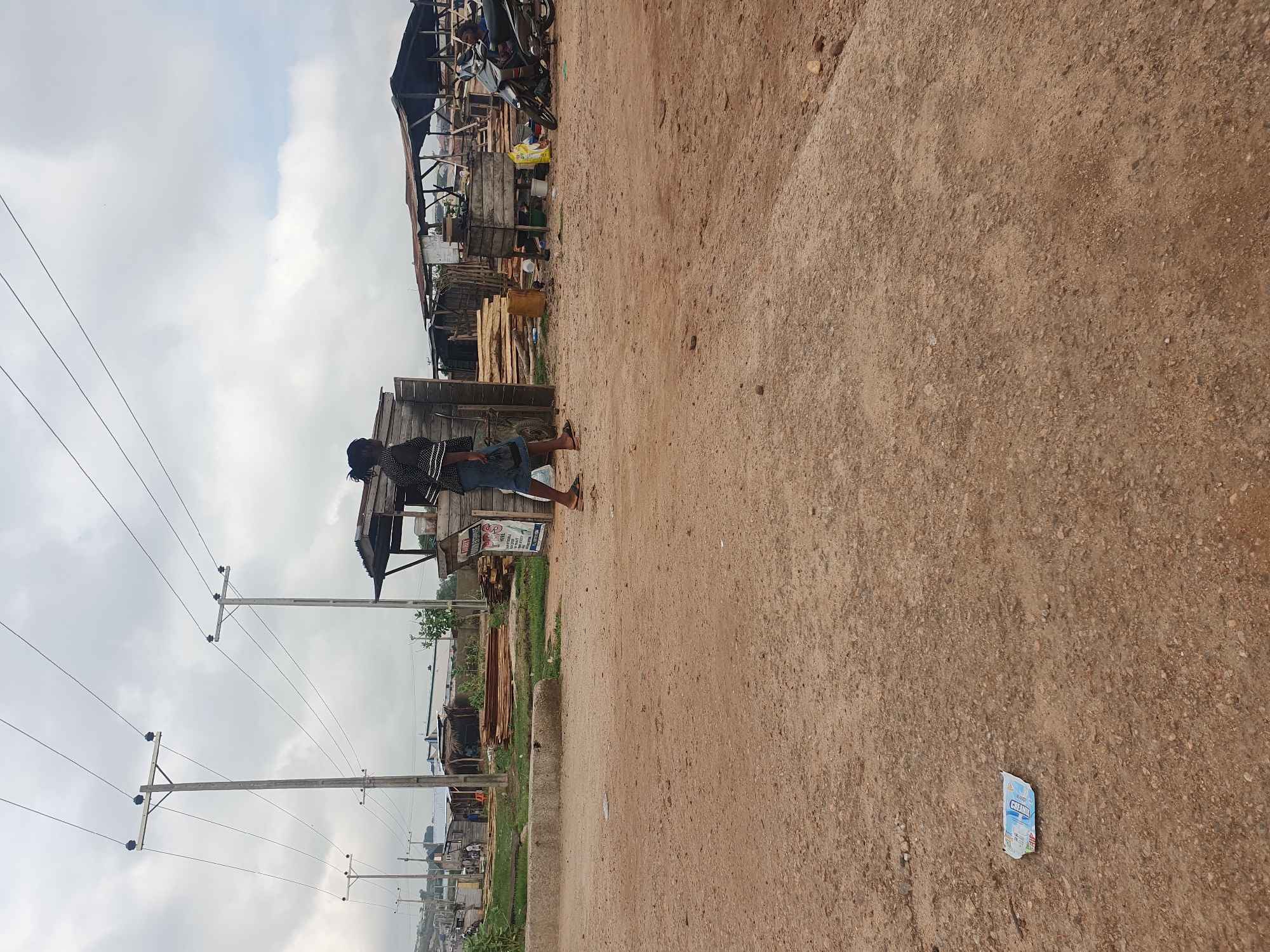 The image depicts a dusty, unpaved road in Lokoja, Nigeria. The road appears to be adjacent to a commercial area with rudimentary wooden structures serving as shops or stalls. Power lines run along the side of the road, supported by concrete poles. A person is visible standing near one of the structures. Debris, possibly a discarded wrapper, lies on the road surface. The sky is overcast.