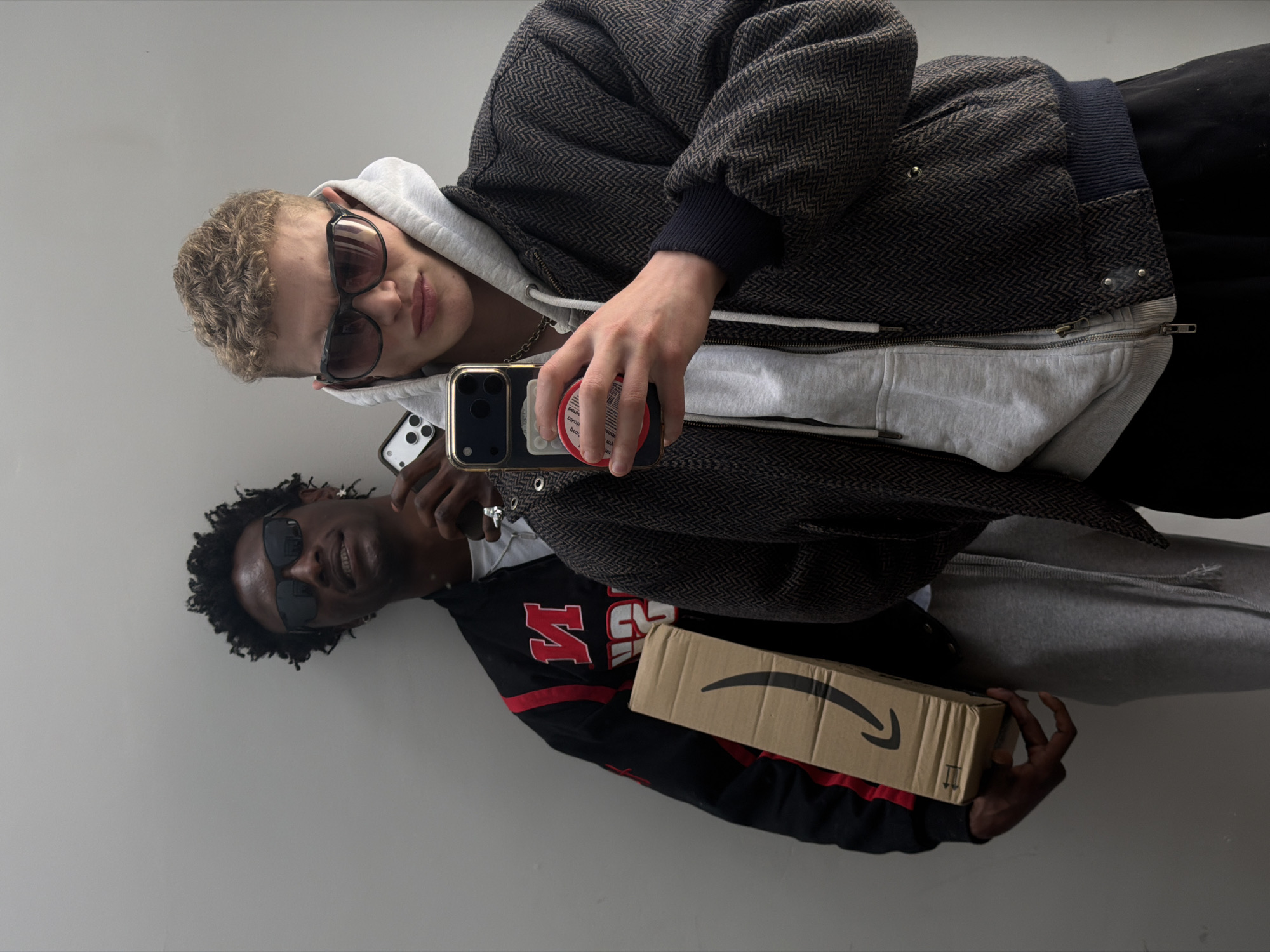 Two people are reflected in a mirror. The individual in the upper portion of the reflection is white, with short, curly blonde hair. They are wearing sunglasses and a light grey hoodie under a dark brown herringbone jacket. The individual is holding a smartphone with a circular pop socket attached to the back. The other person is Black and wearing sunglasses and a black sweatshirt with red accents. The person is holding a cardboard box with the Amazon logo printed on it. The background is a plain white wall. There is no indication of the location being in London.