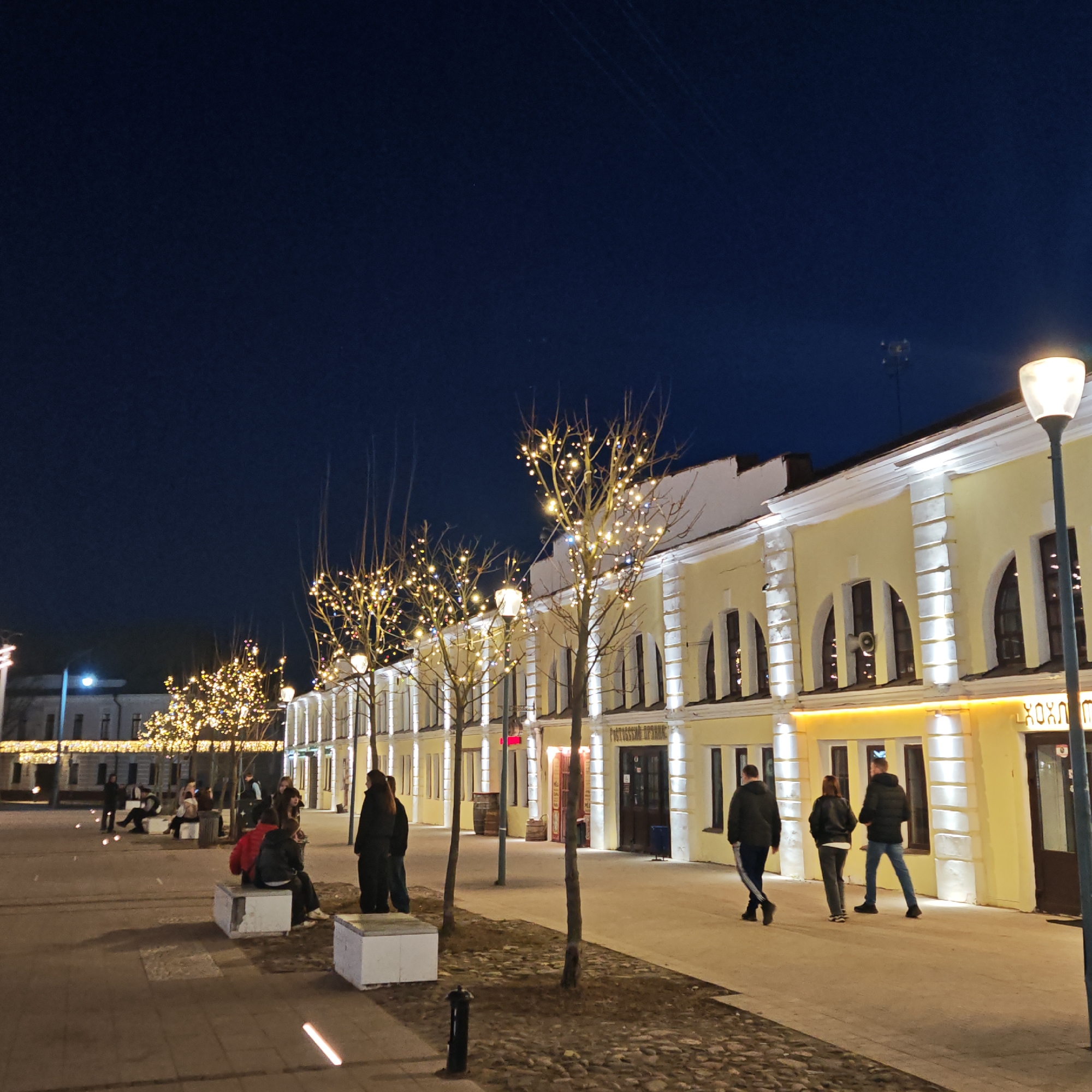 The image depicts a nighttime scene in Rostov, Russia, featuring a line of yellow buildings with arched windows and decorative lights strung on trees in front. People are seen walking along the sidewalk and sitting on benches. Streetlights illuminate the area. The sky is a deep blue, suggesting it is dusk or later. A cobblestone strip runs parallel to the sidewalk. The buildings appear to be commercial establishments.
