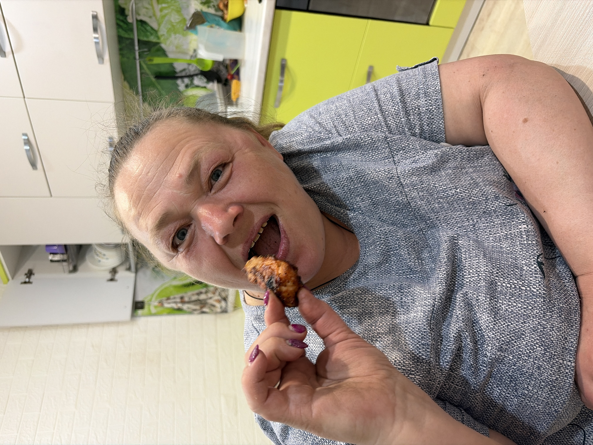 A Caucasian woman is in a kitchen, holding a cooked food item between her thumb and index finger. She is in the process of putting the food into her mouth. The location is a kitchen with white and lime green cabinets. The woman has gray eyes, short light brown hair, and is wearing a gray and white shirt. Her fingernails are painted with a sparkly purple polish. The environment includes a white textured backsplash and white cabinets with silver handles. The location details of being in Noviy, Russia is not evident.