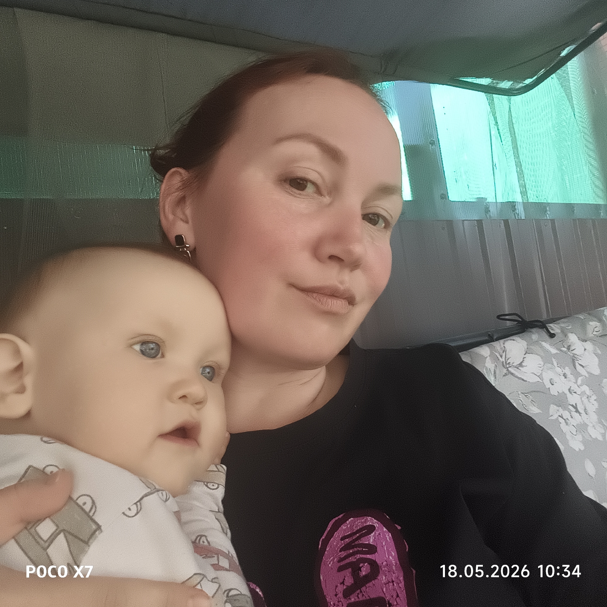 A woman is holding a baby. The woman has fair skin, reddish-brown hair pulled back, and is wearing a black long-sleeved shirt. The baby has fair skin and blue eyes. The photo appears to be taken indoors, with a translucent green material visible in the background. The date and time "18.05.2026 10:34" are stamped in the bottom right corner, along with the camera information "POCO X7" on the bottom left. There are cushions with a floral pattern in the background. The location of the image is presumed to be Chaikovsky, Russia.