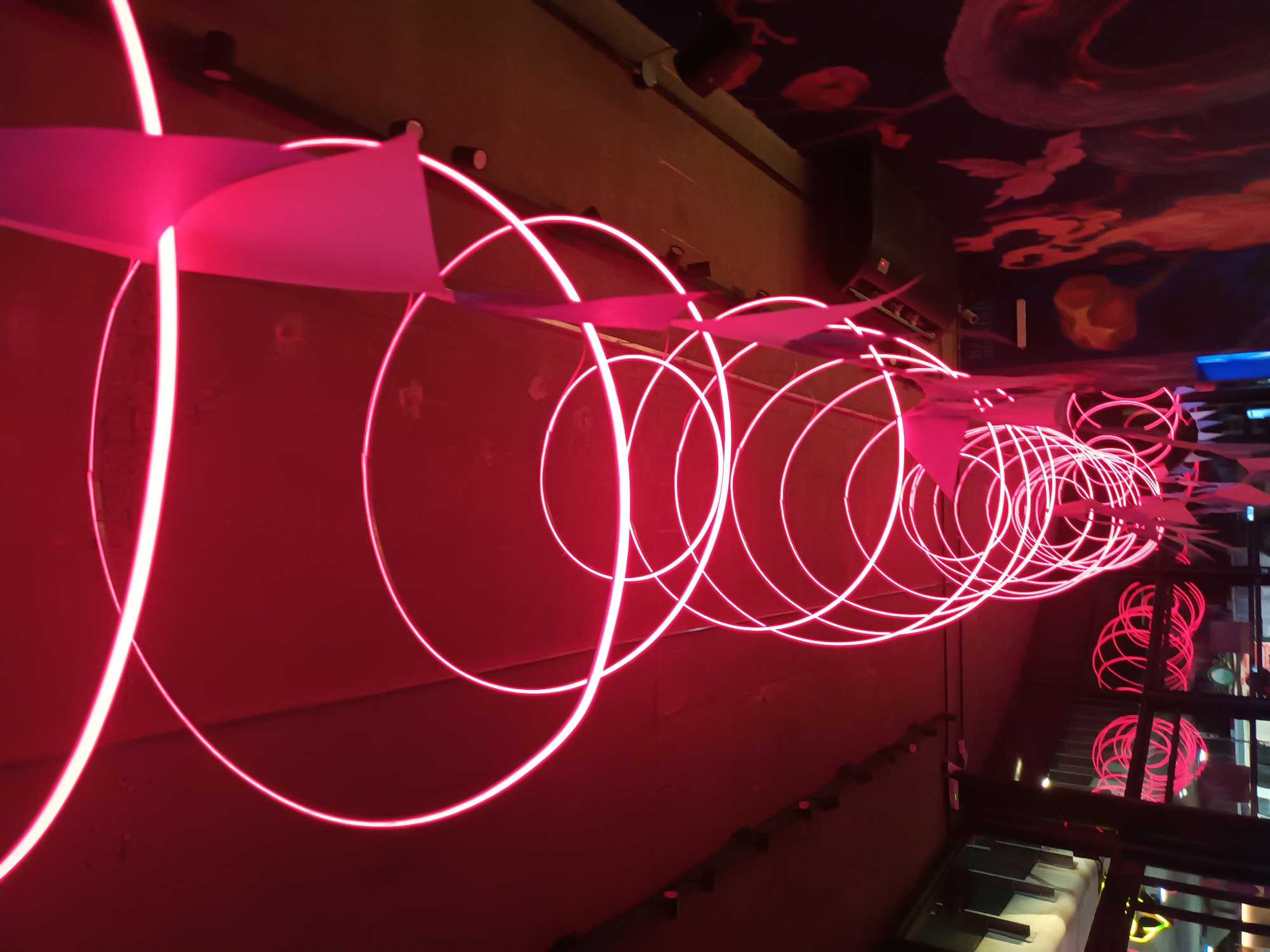 A high-angle shot captures an indoor scene featuring a series of neon pink circles suspended from the ceiling. The circles decrease in size as they recede into the background, creating a sense of depth. Pink paper decorations are interspersed among the neon circles. The walls are painted in a matching shade of pink. The ceiling is decorated with a mural. In the background, a window provides a glimpse of the outdoors and a piano. The location is Buenos Aires, Argentina.