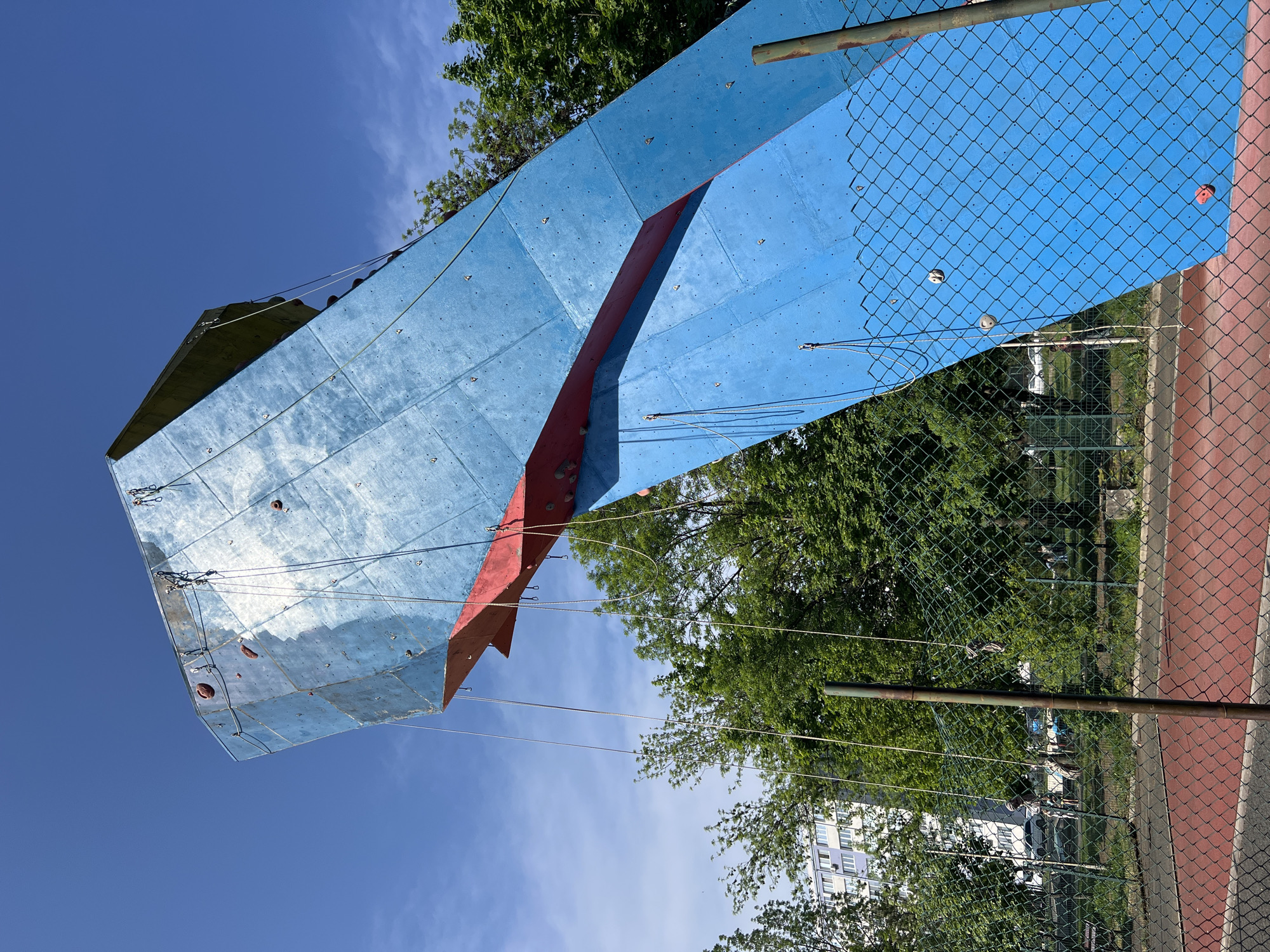 The image shows a blue and red outdoor climbing wall structure in Burgas, Bulgaria. The climbing wall is next to a green chain-link fence with a red base. The sky is blue with scattered clouds, visible behind and reflected in the climbing wall's surface. Green trees are present in the background. In the distance, a light-colored apartment building is visible among the trees.