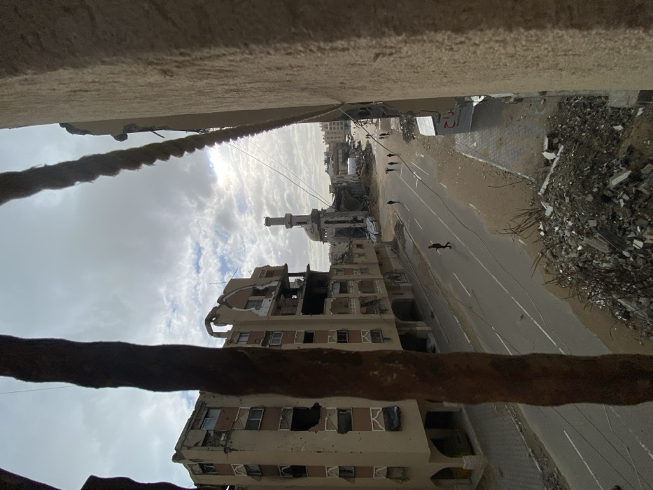 A low-angle shot shows severe destruction; a beige building with shattered windows and structural damage stands prominently. A minaret rises from the wreckage nearby. The street below is littered with debris, contrasting sharply with a line of figures walking away. Overhead, a cloudy sky dominates the frame, while blurred elements of the structure from which the photo is taken frame the top and bottom.