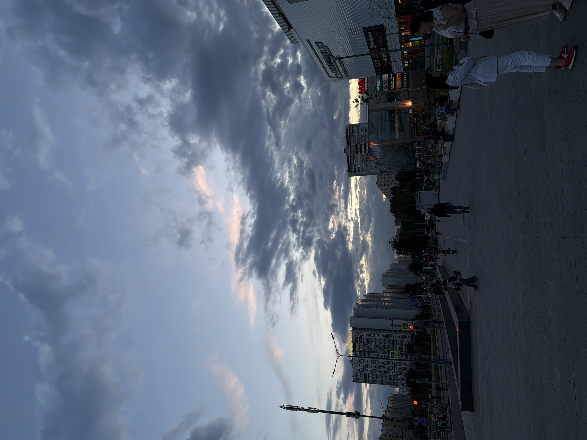 The image depicts an urban street scene or plaza in Moscow, Russia, captured during late afternoon or early evening. The sky is a prominent feature, dominated by dark grey, voluminous clouds, interspersed with patches of lighter blue and subtle streaks of pink and orange, hinting at a sunset or recent weather transition.
Several people are present in the scene, engaging in casual public activity. In the right foreground, two women are visible: one in white attire with red sneakers, and another in a light-colored pleated skirt and top. Other individuals are scattered across the paved ground, walking or standing.
To the right, a large commercial building features multiple storefronts with visible Cyrillic signage. These include "MAX", "Пятерочка" (Pyaterochka, a supermarket), "Перекресток" (Perekrestok, another supermarket), "Талавера", "OFFPRICE", "ОПТИКА" (Optika, an optician), and "СУШИ" (Sushi). This indicates a busy commercial district. In the midground and background, residential high-rise buildings rise under the cloudy sky.
The ground consists of a wide, paved area, suggesting a pedestrian zone. Urban infrastructure like streetlights, a red traffic light, and a modern light pole or cell tower are also visible. The overall impression is that of a contemporary city environment as day transitions into dusk.