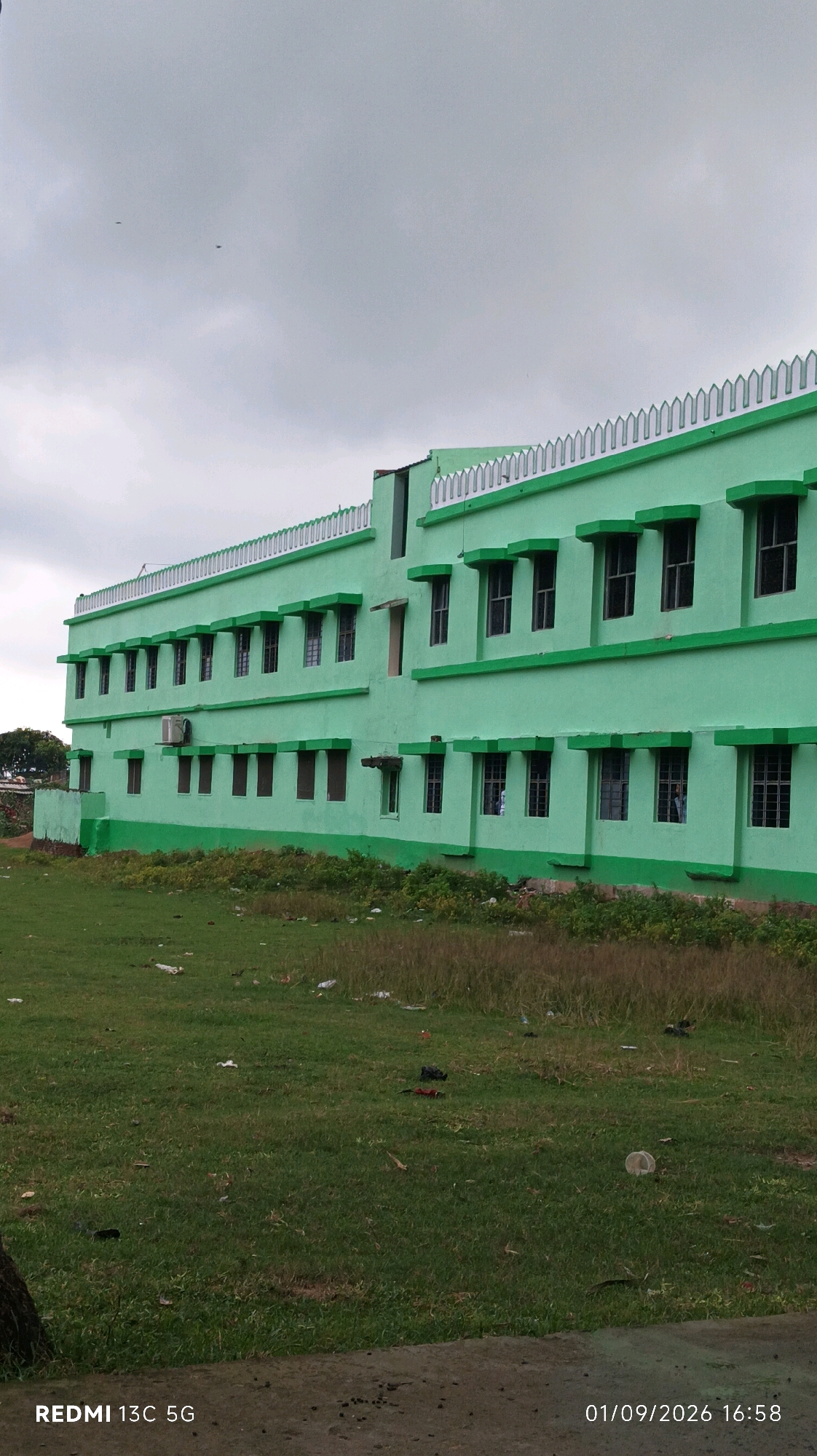An exterior, full-shot, eye-level, vertical, medium-distance photograph captures a three-story, light-green building in India under a cloudy, gray sky. The building's facade is uniform, with rows of identical windows on each floor, each featuring a dark-green awning-like structure above. The building's roofline is adorned with a decorative white, crenellated border. In front of the building, a green, grassy field is visible, littered with various debris including plastic bottles, wrappers, and some darker objects. The field extends to the bottom of the frame. The date and time stamp "01/09/2026 16:58" and the text "REDMI 13C 5G" are present in the lower right and left corners of the image, respectively. The scene appears to be an outdoor educational institution or a similar public building.